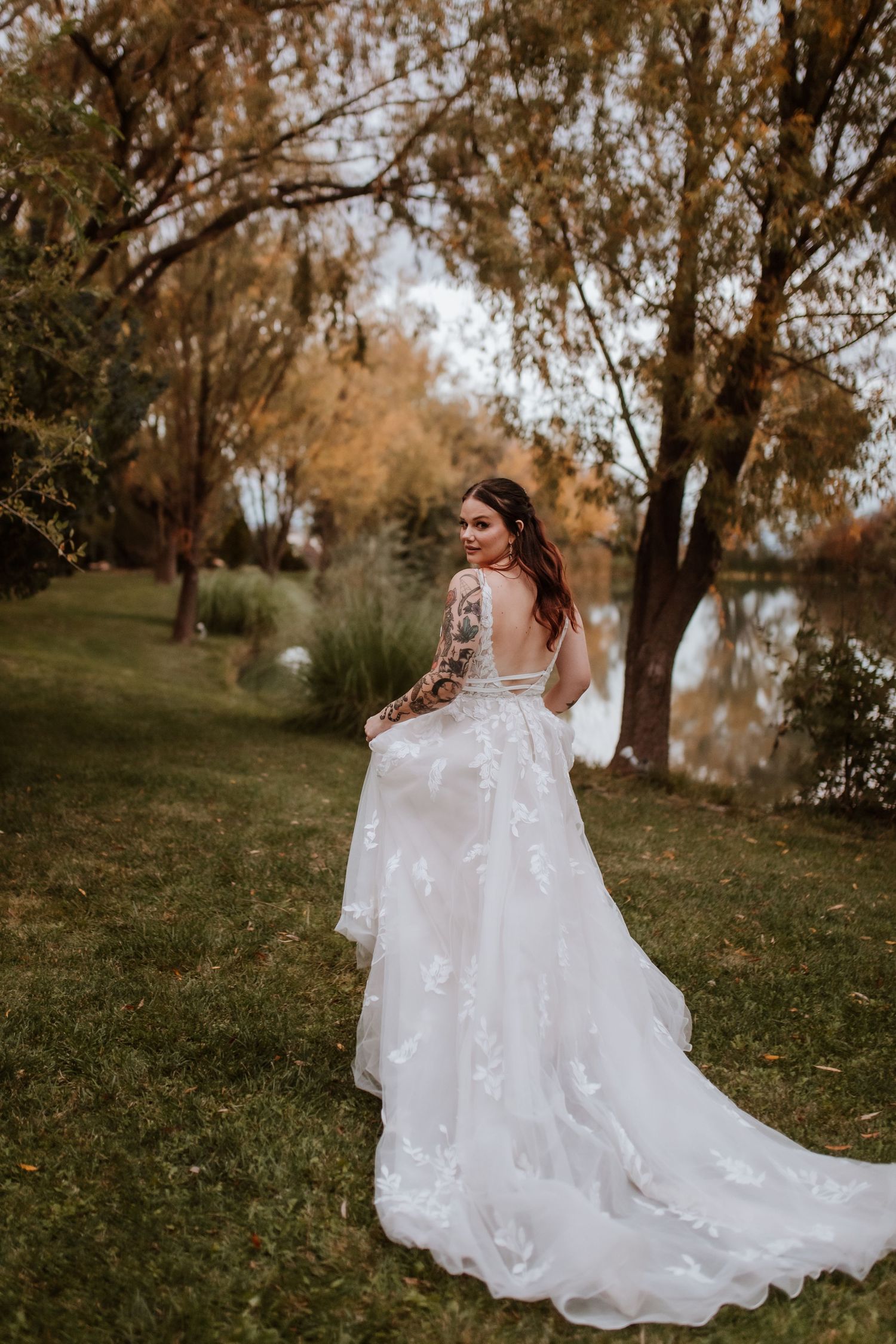 A wedding dress with lace details and flowing train photographed outdoors against autumn trees near a pond by tucson wedding photographer