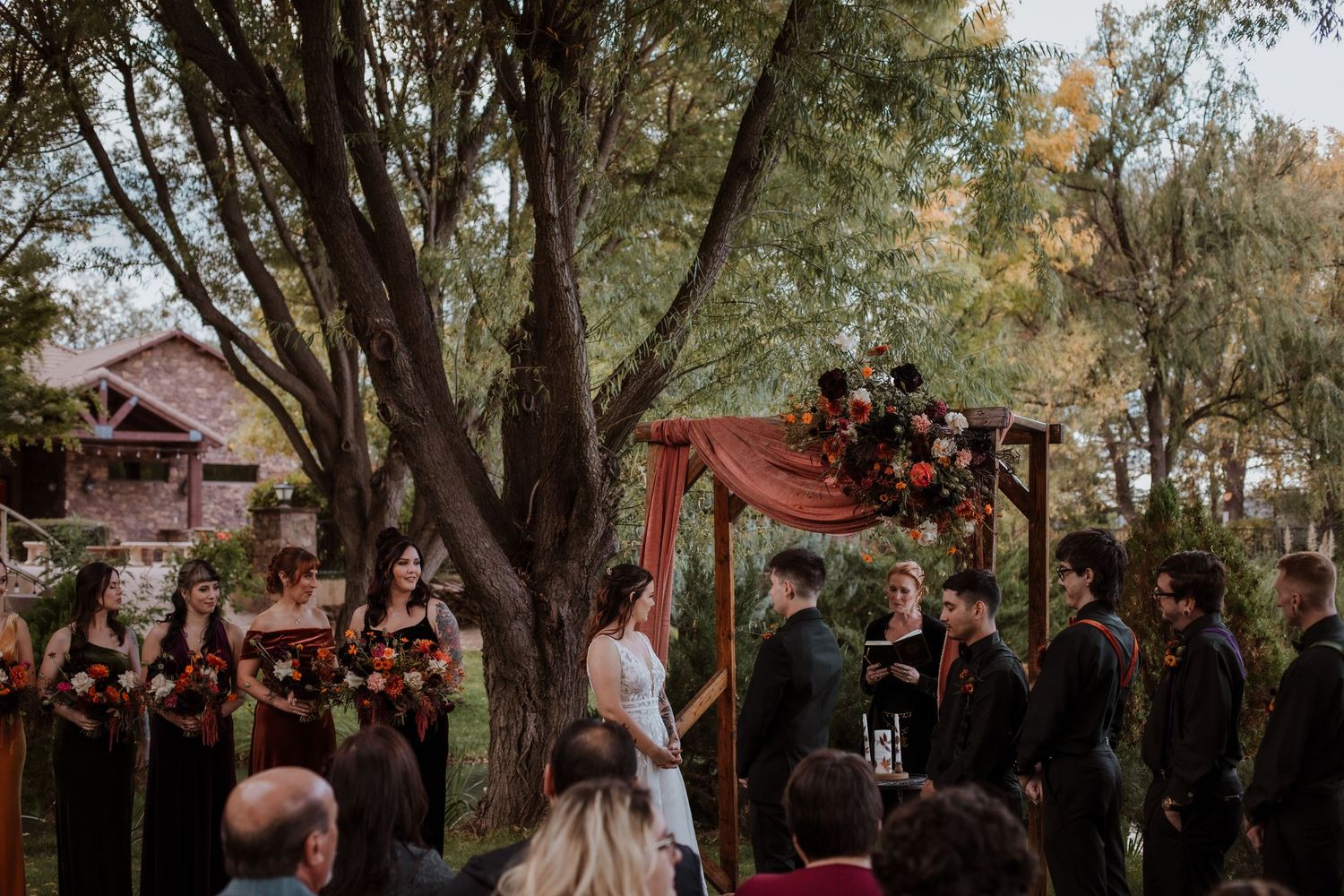 A wedding ceremony takes place under a large tree with a wooden arch and burgundy draping. by sierra vista wedding and boudoir photographer