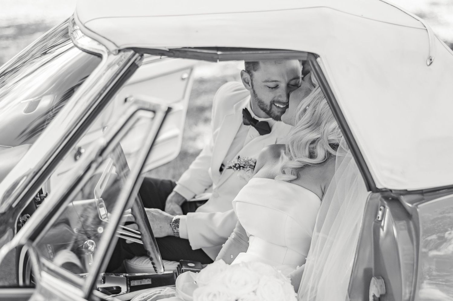 A black and white shot of a couple sitting in a classic, vintage car learning in for a kiss on their wedding day