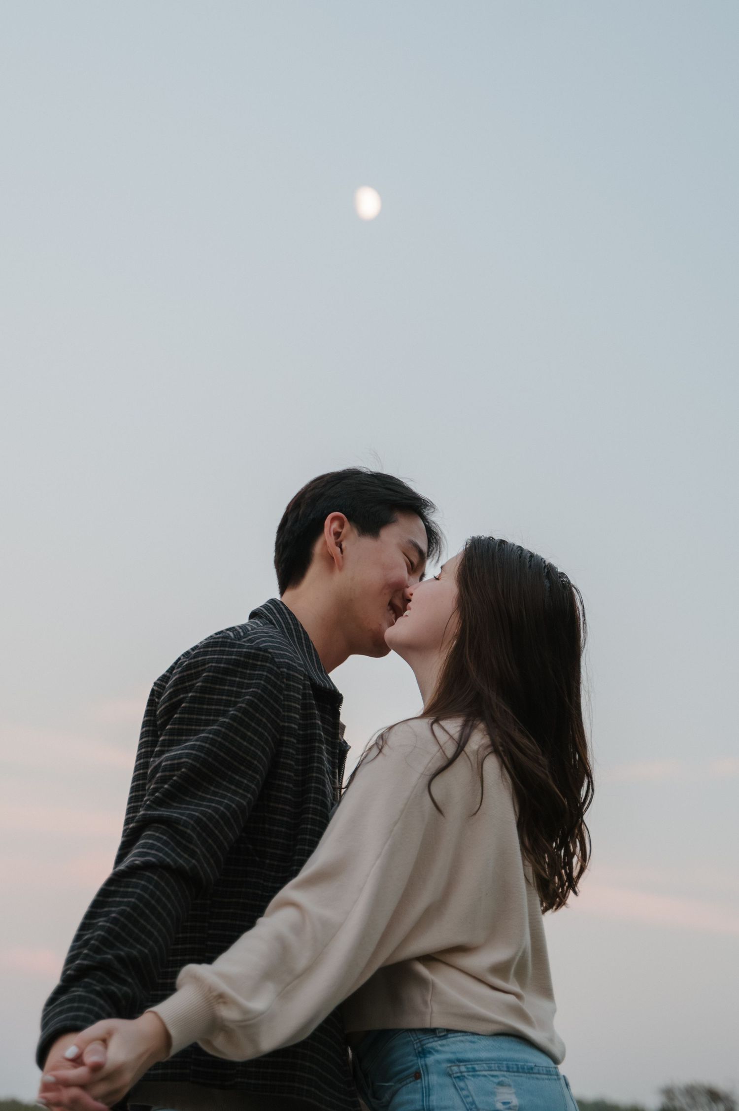A couple embracing and leaning in for a kiss as the sun sets with cotton candy pink clouds on the horizon and a half moon in an icy blue sky