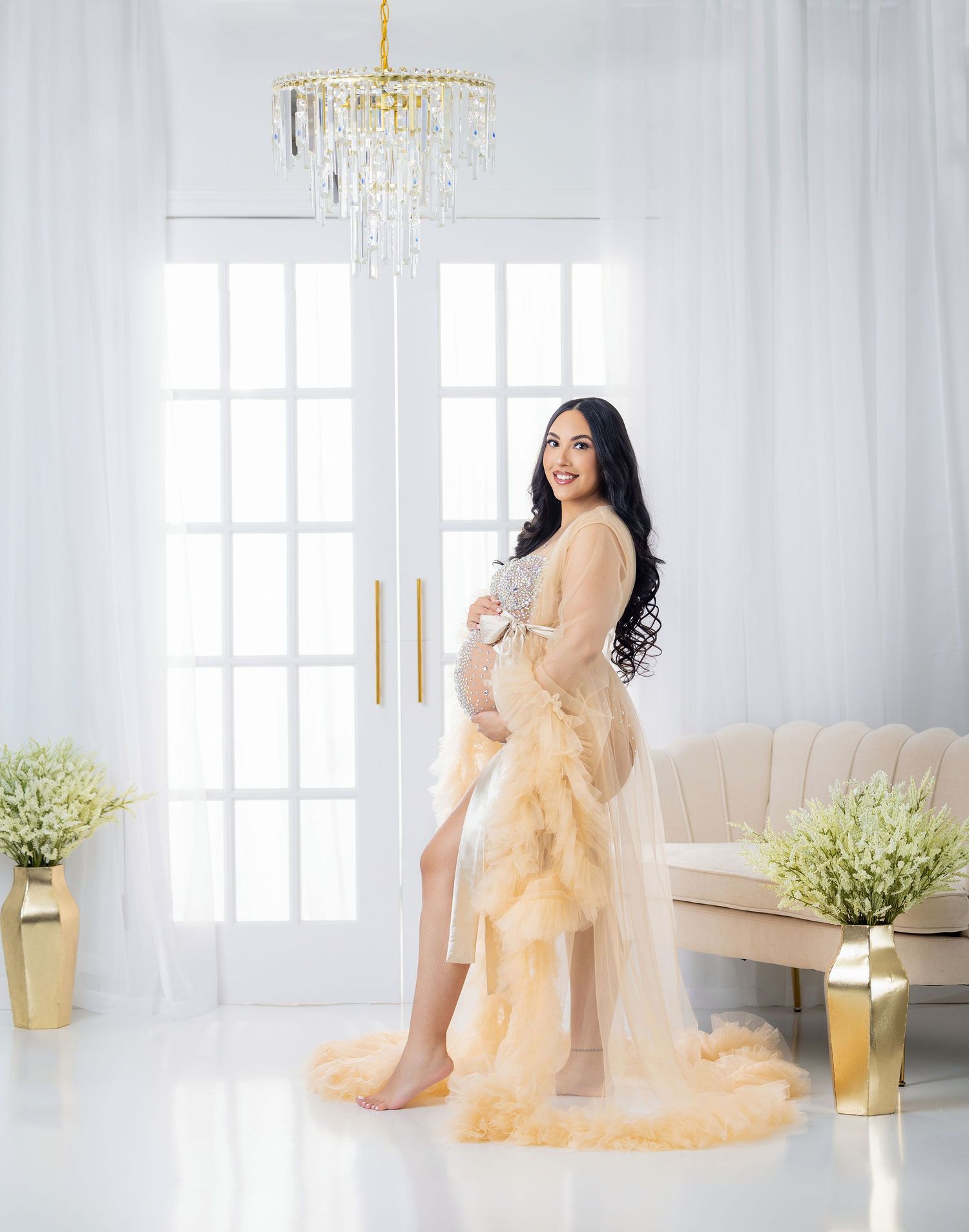 Elegant maternity photo in white room with crystal chandelier and gold accents featuring flowing champagne tulle gown.