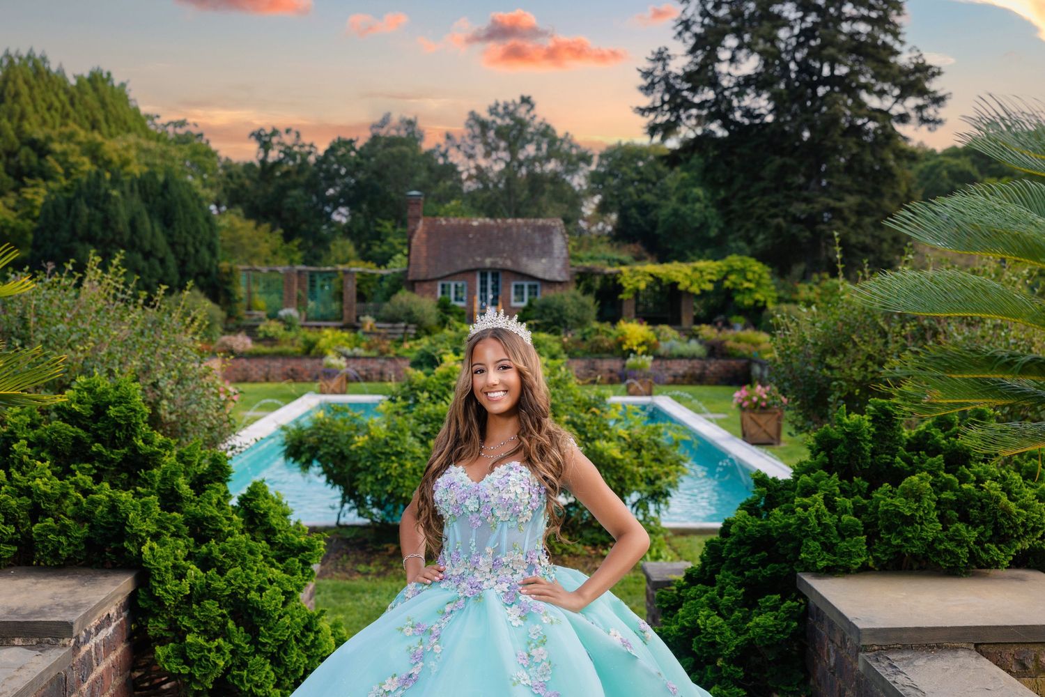A person in a light blue ballgown poses in a garden setting with a swimming pool and cottage at sunset.