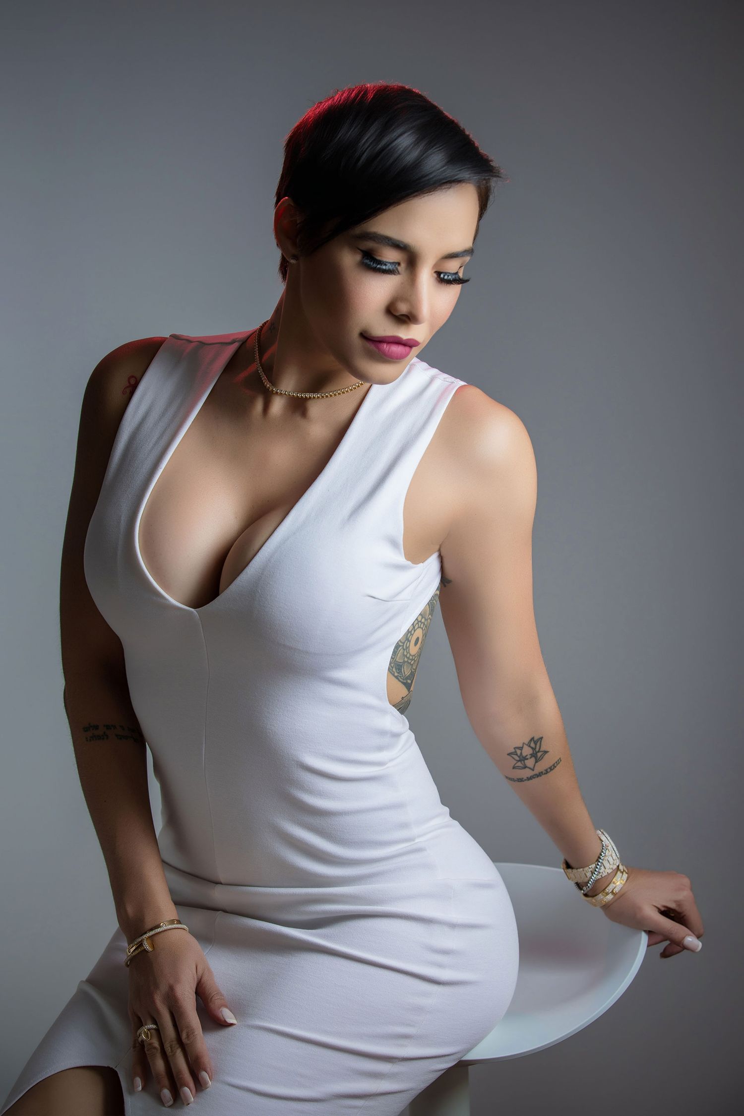 Woman in white deep-V dress with tattoos posing gracefully in gray studio setting.