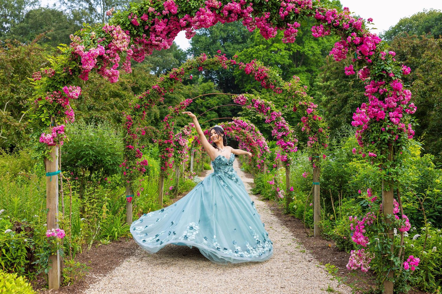 A figure in a flowing blue ballgown poses gracefully among vibrant pink climbing roses in a garden pathway.