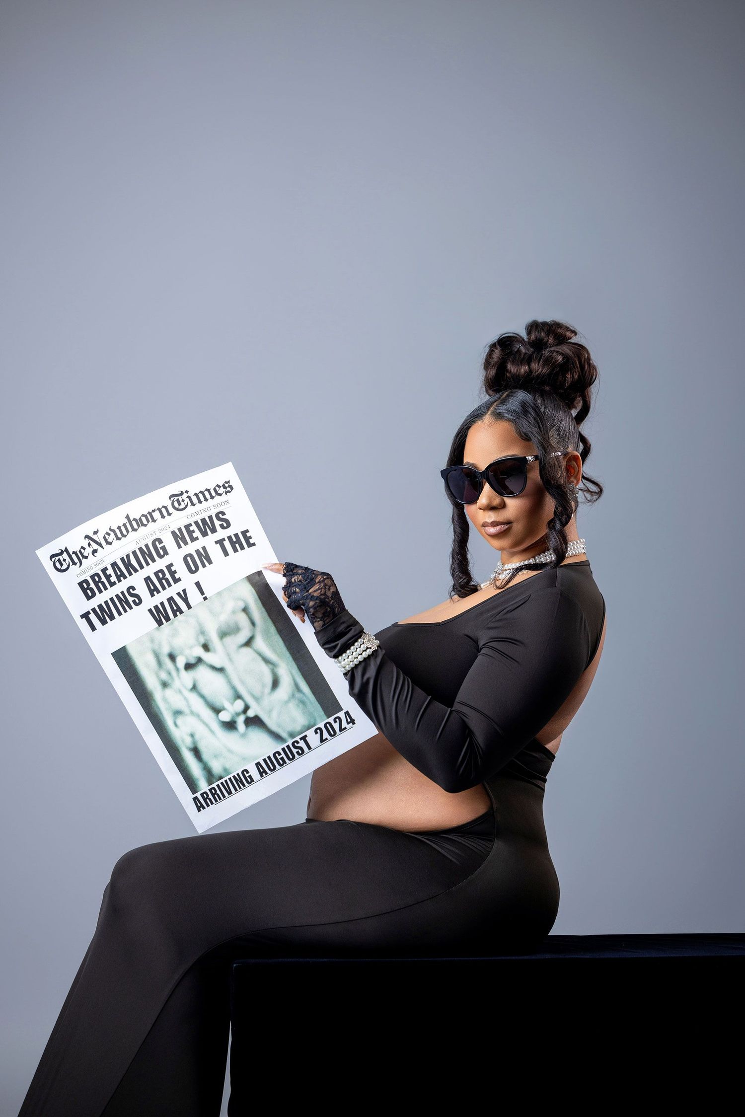 A person in black attire reclines while reading a magazine against a gray studio backdrop.