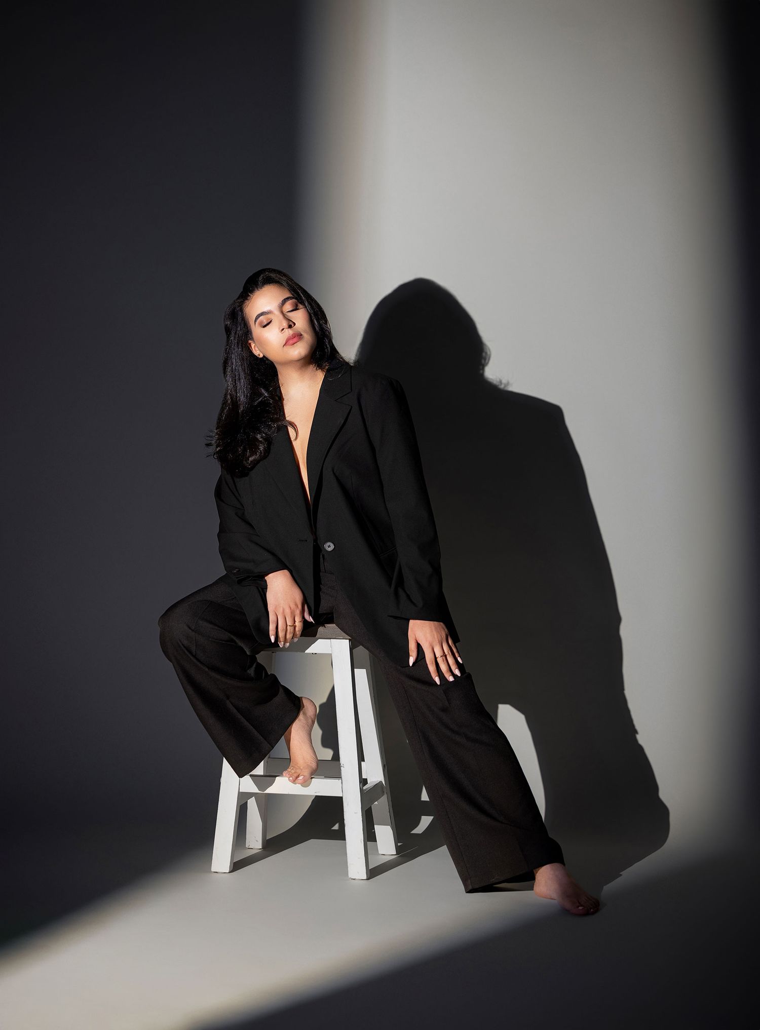 Woman in black oversized blazer and wide-leg pants seated on white stool with dramatic shadow.