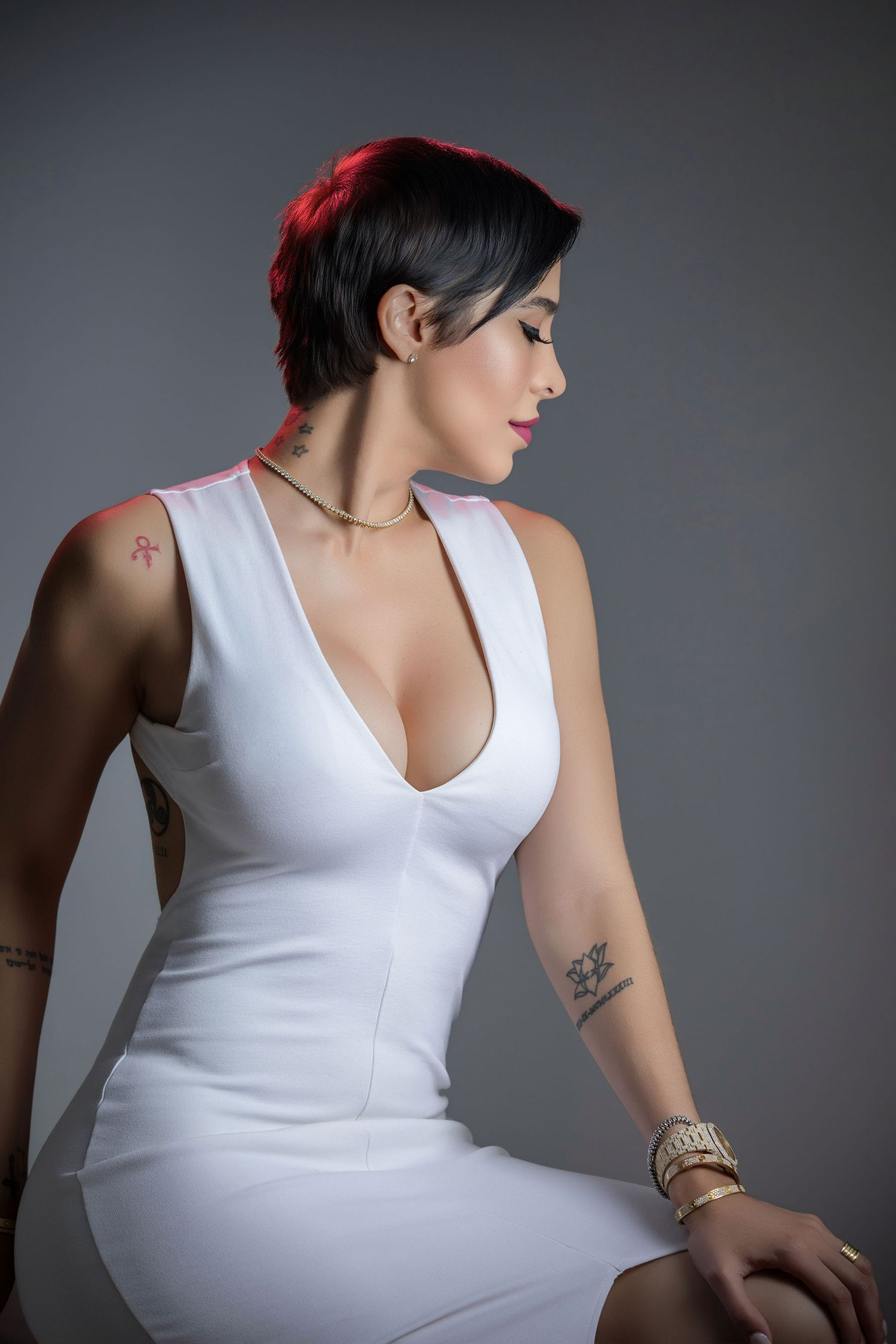 Woman with short hair in white sleeveless dress with tattoos poses in studio.
