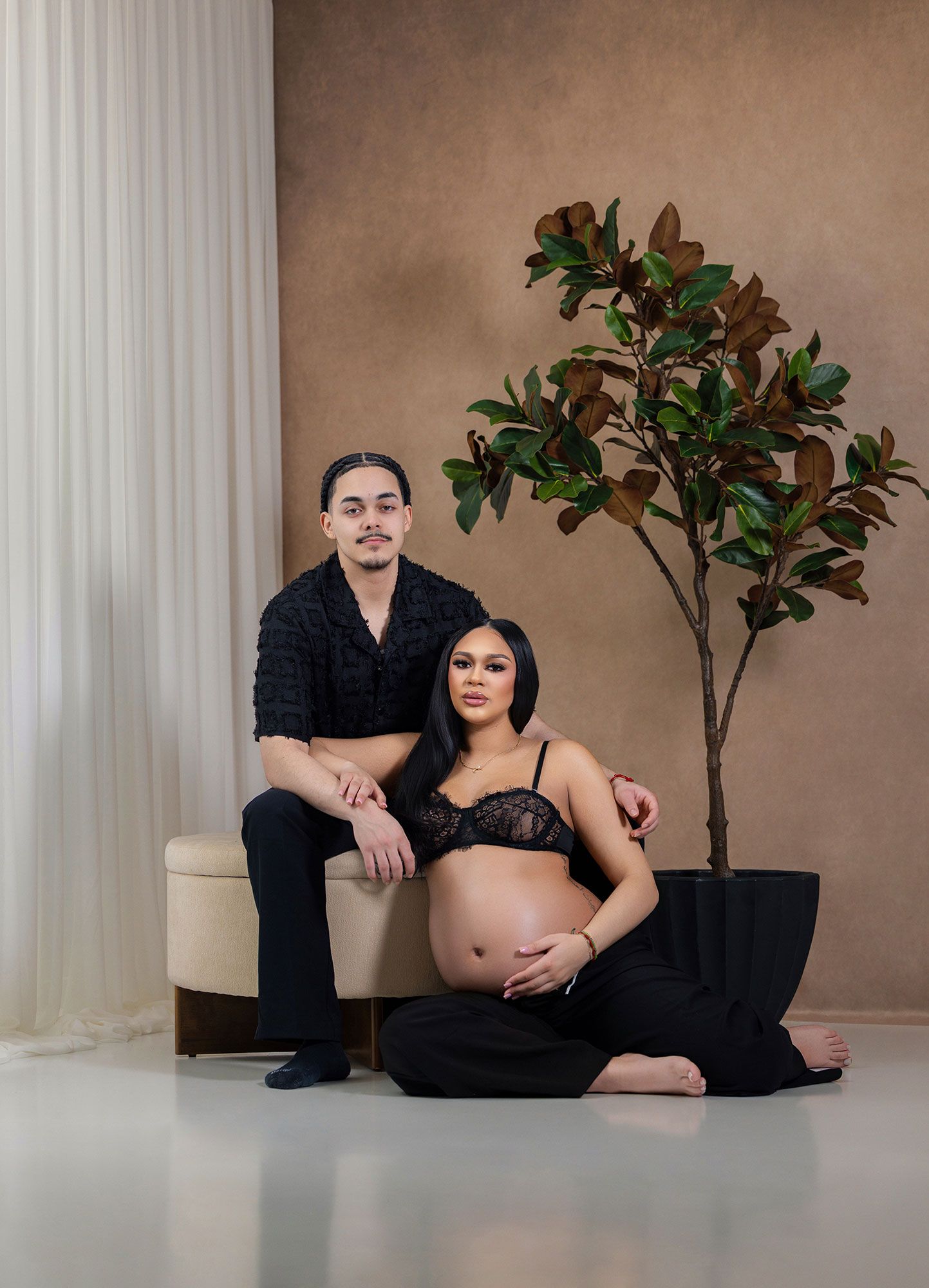 A couple poses in an artistic maternity photoshoot with white columns and fiddle leaf fig plants in the background.