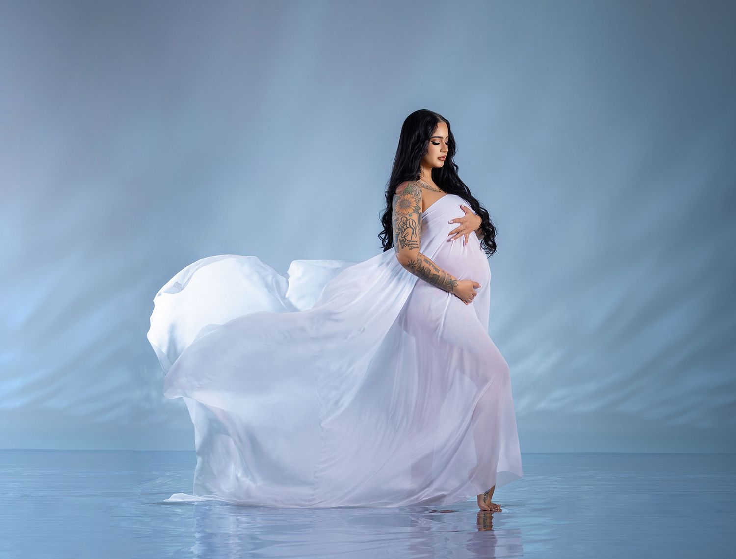 A flowing white dress billows gracefully in a soft blue-toned studio setting creating an ethereal artistic effect.