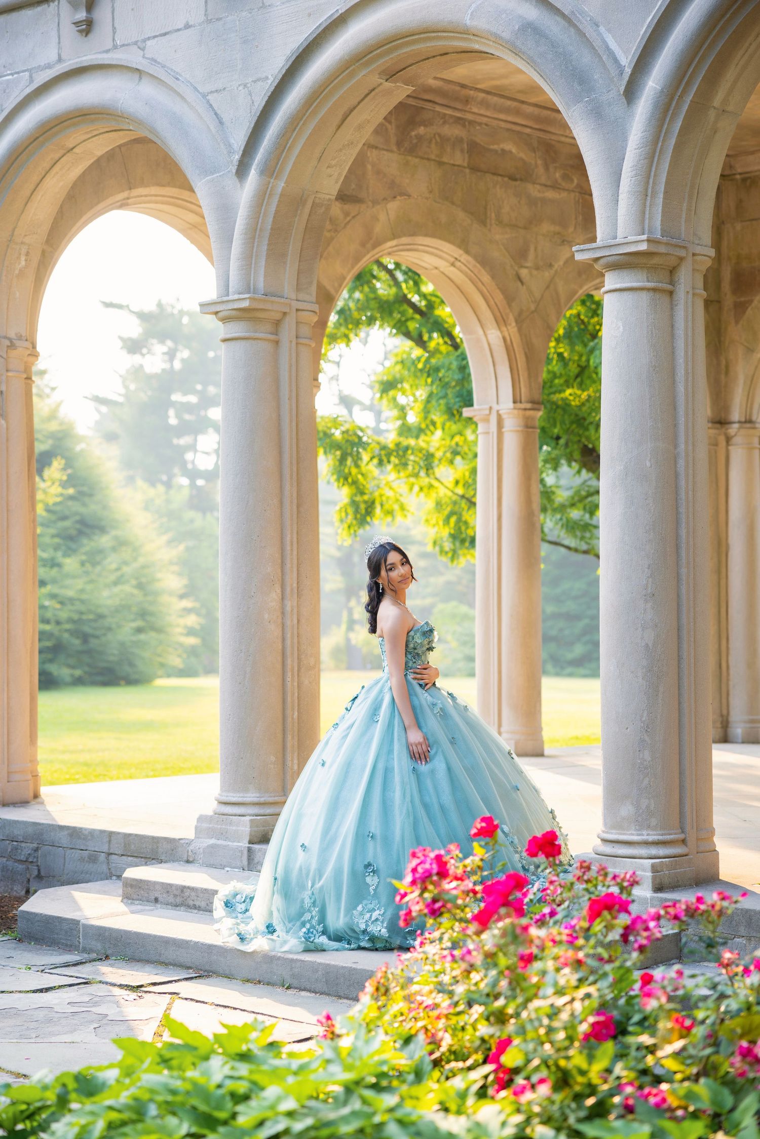 A woman in a light blue ball gown poses under stone arches surrounded by lush greenery and red flowers.