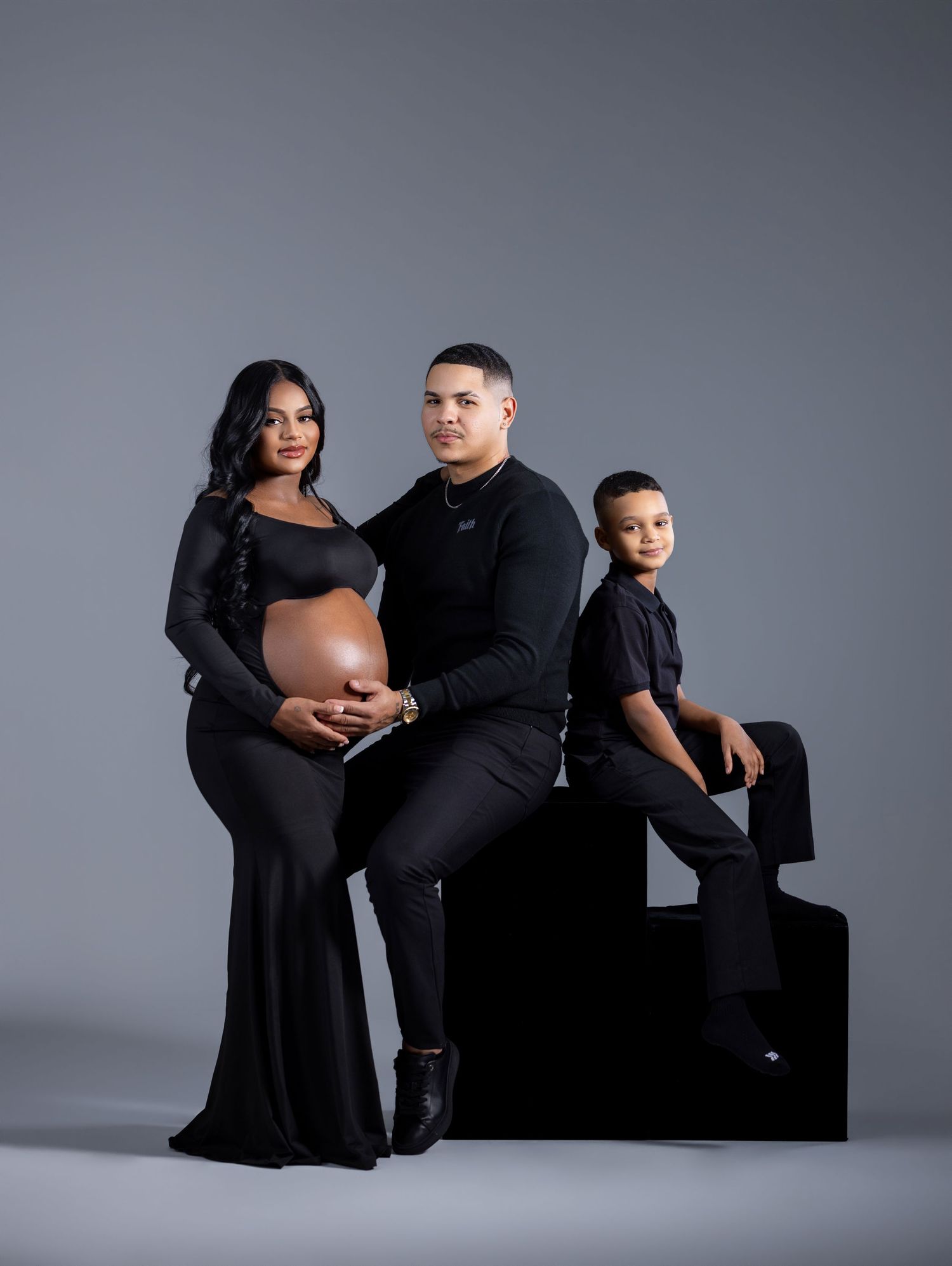 Elegant maternity photo of a pregnant woman in black gown with partner and child posing in studio.
