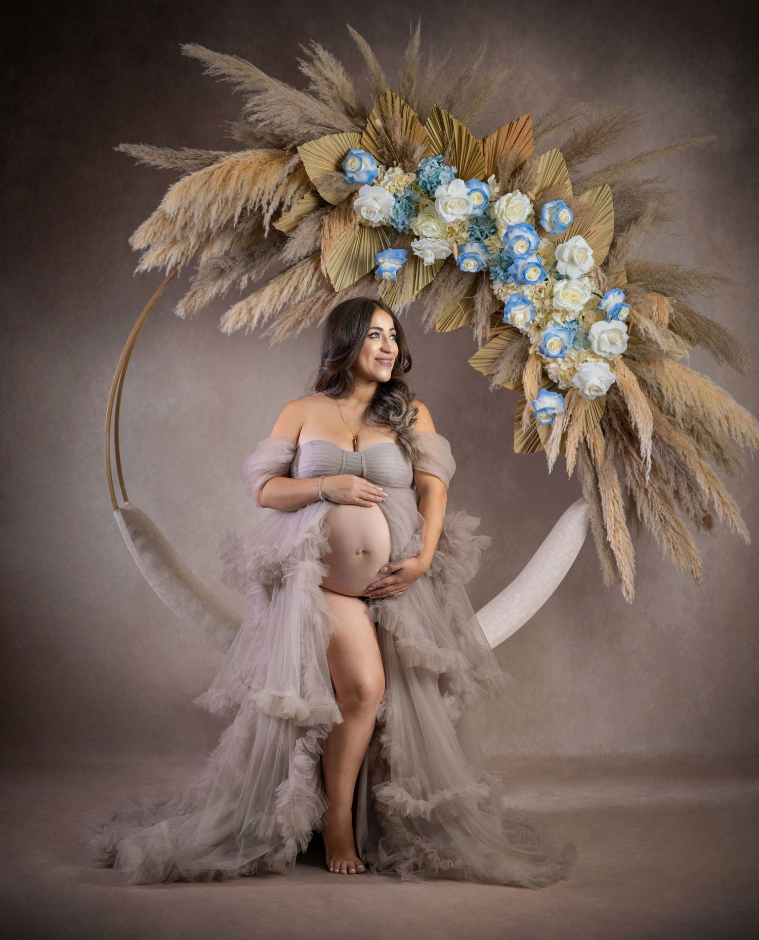 Maternity photo featuring flowing gray tulle dress and floral arch with pampas grass and blue roses in studio setting.