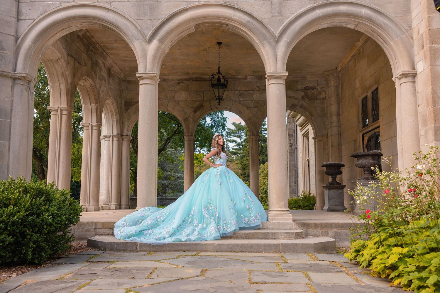 Young woman in a flowing teal quinceañera ball gown poses gracefully under stone arches of an elegant mansion.