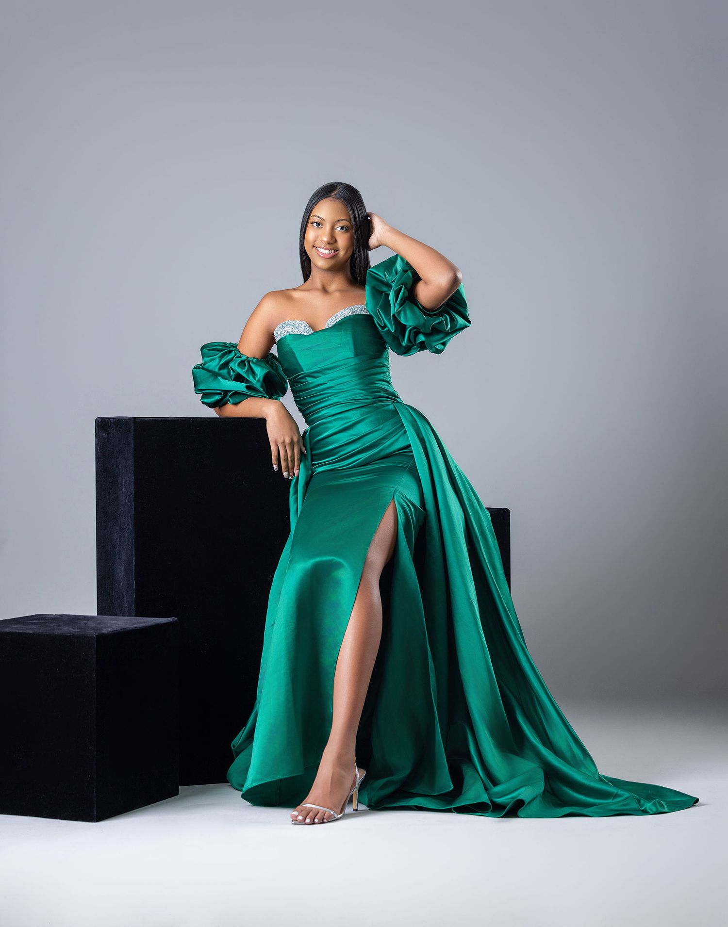 Woman in elegant emerald green off-shoulder satin gown with slit posing in a studio.