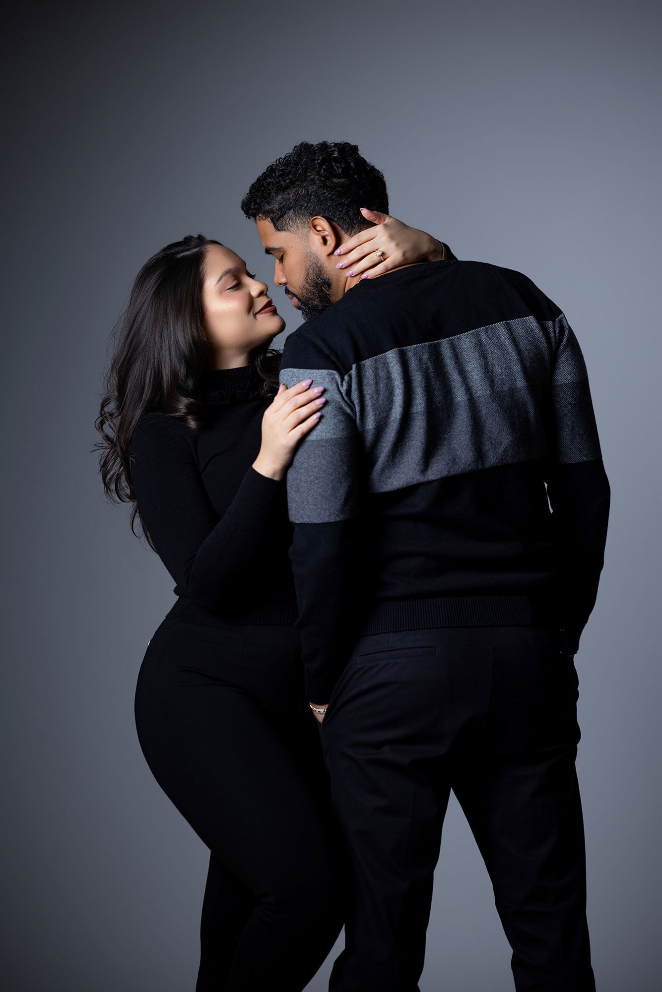 A couple in dark clothing embraces intimately against a gray studio backdrop in a series of romantic poses.
