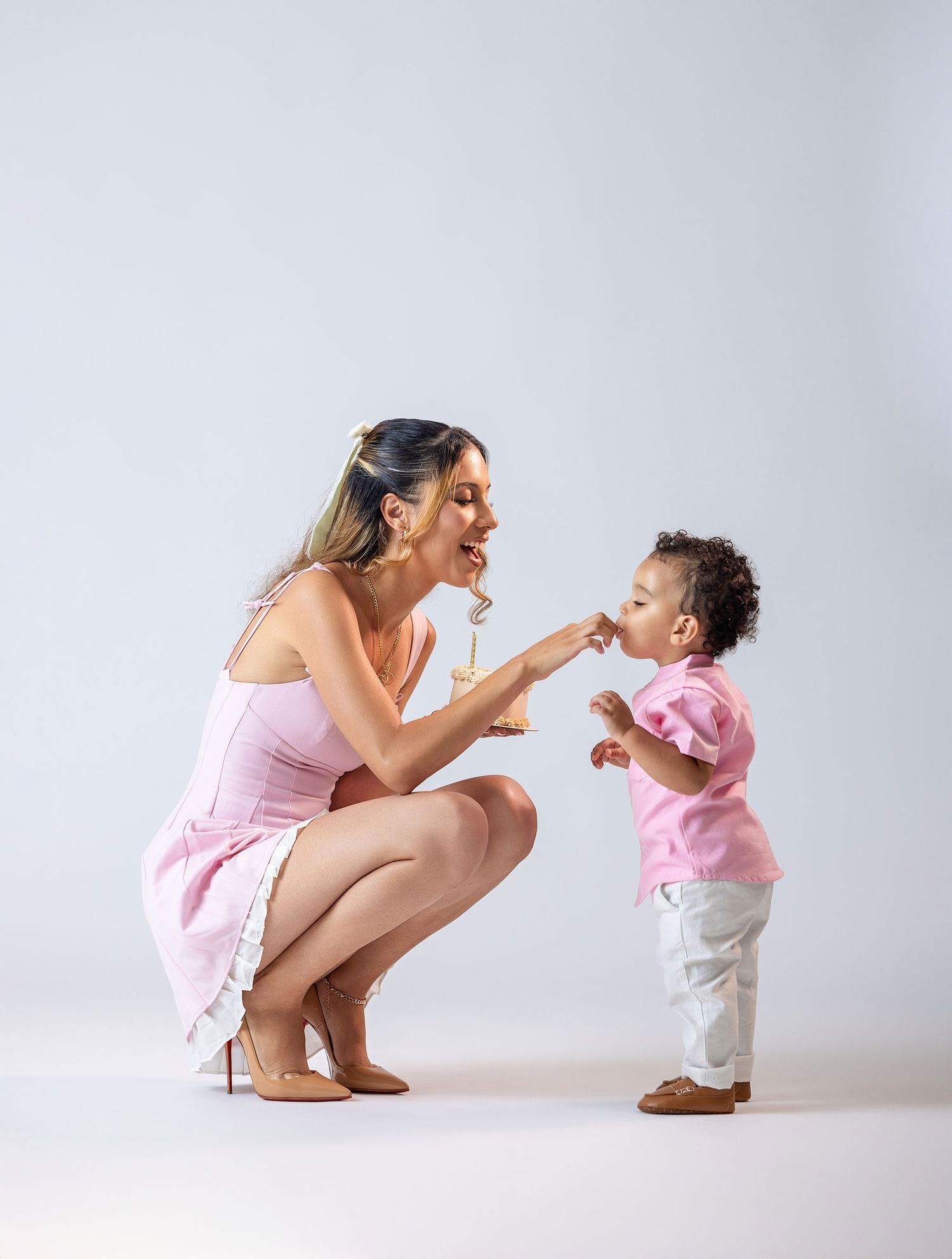 Woman in pink dress crouching to interact with toddler in white studio setting.