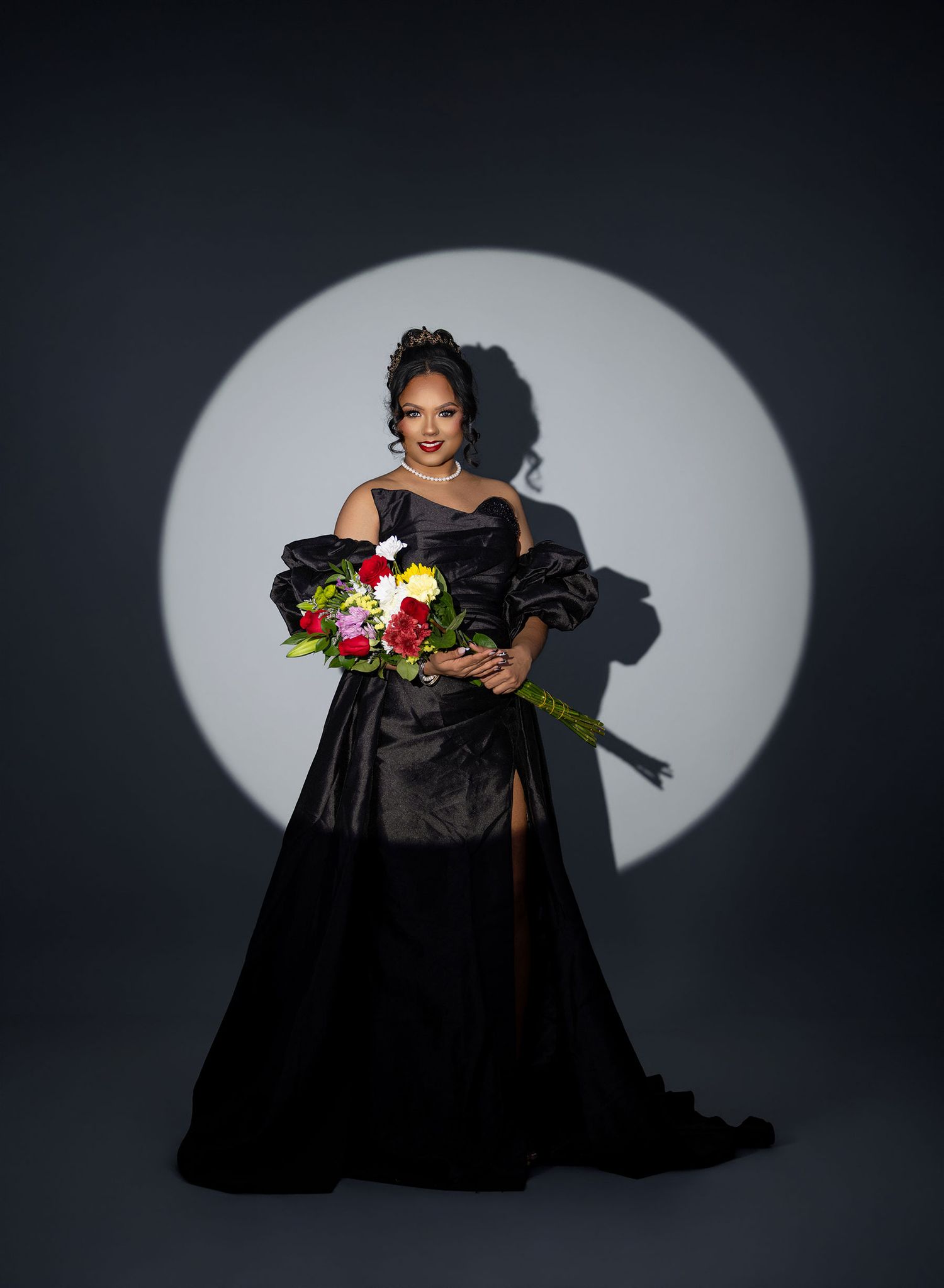 Woman in elegant black off-shoulder gown holding colorful bouquet, posed against circular spotlight backdrop.