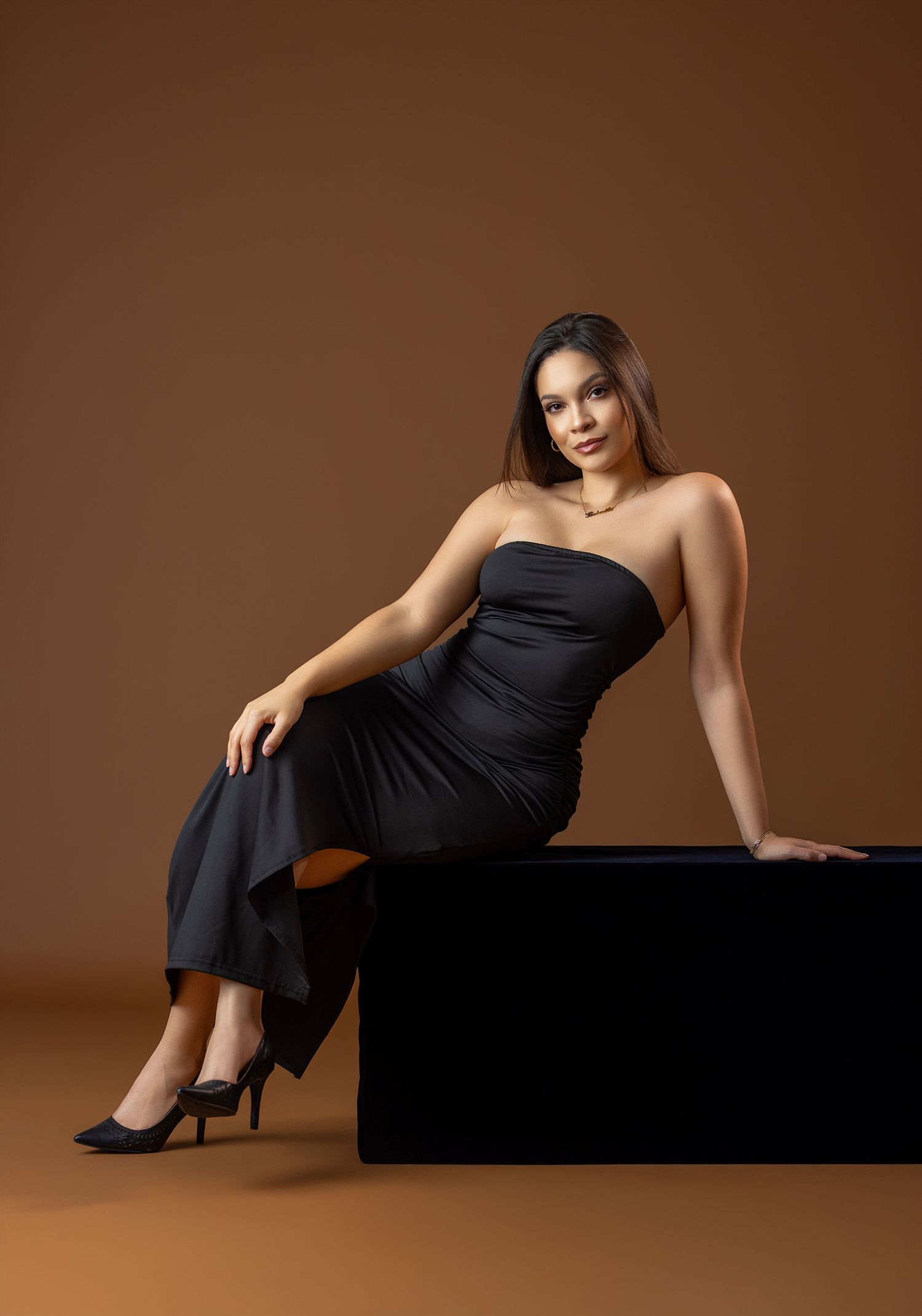 Woman in black strapless gown seated on black cube against warm brown background.