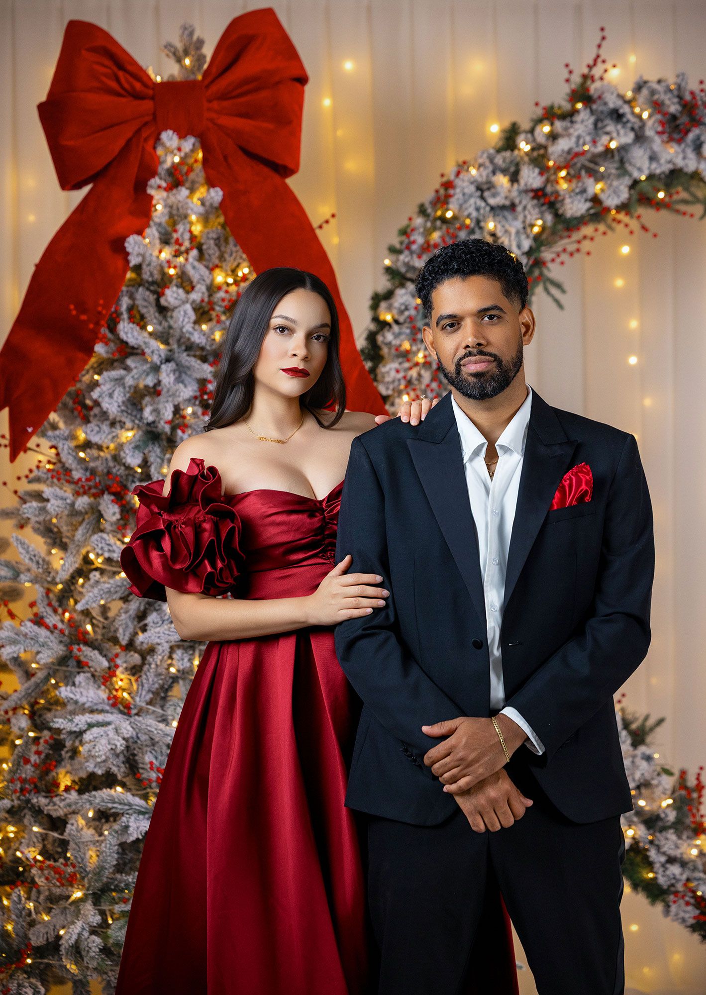 Elegant couple poses in front of silver Christmas tree with red bows and twinkling lights in formal holiday attire.