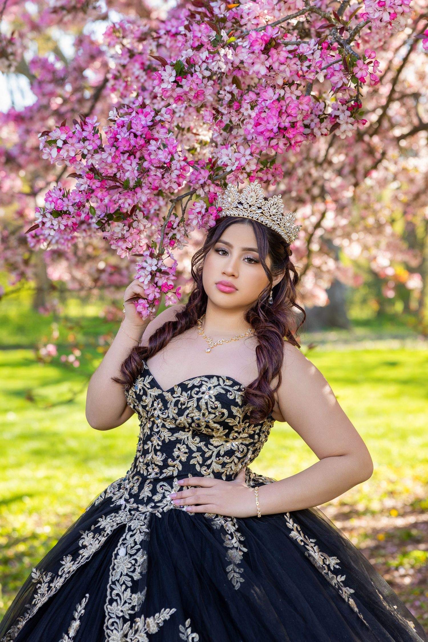 Young woman in black and gold quinceañera gown and crown poses before pink cherry blossom tree.