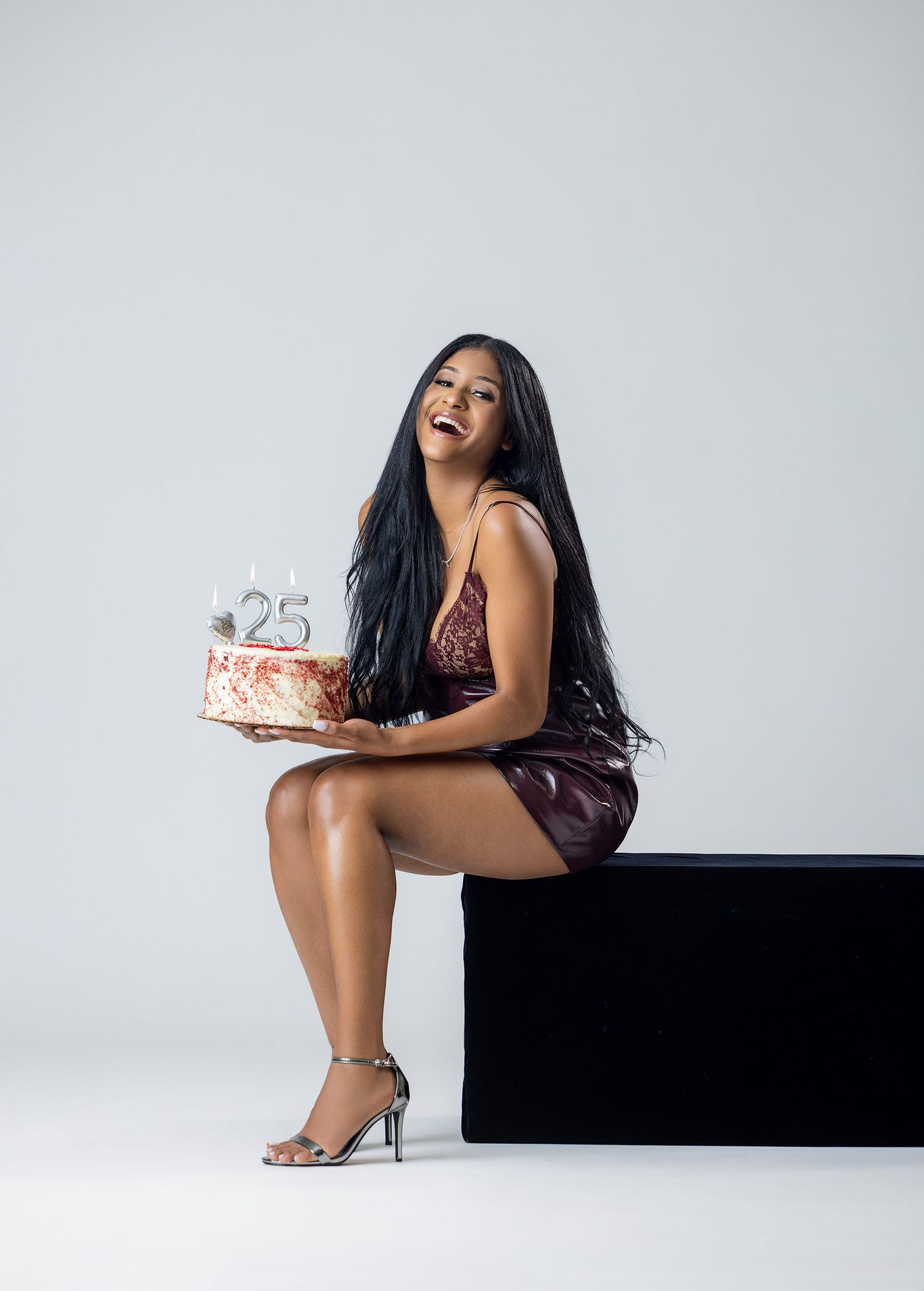 Woman in burgundy dress holding a birthday cake with number 25 candles in a studio.