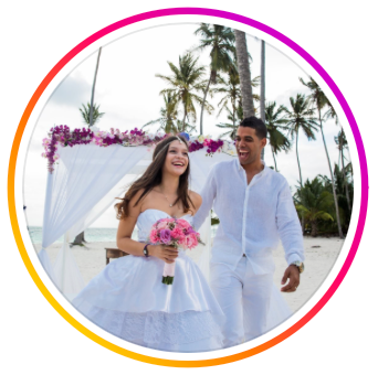 Couple in white attire walking along a tropical beach with palm trees during sunset celebration.