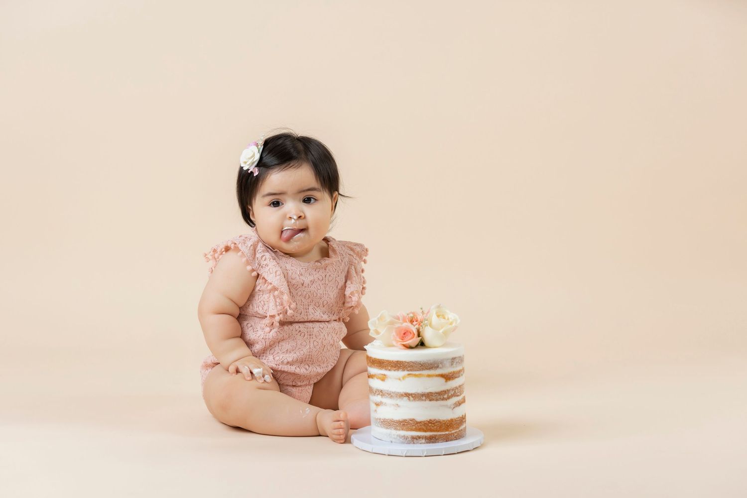 Baby girl in pink romper sits beside a naked birthday cake during a first birthday cake smash photo session.