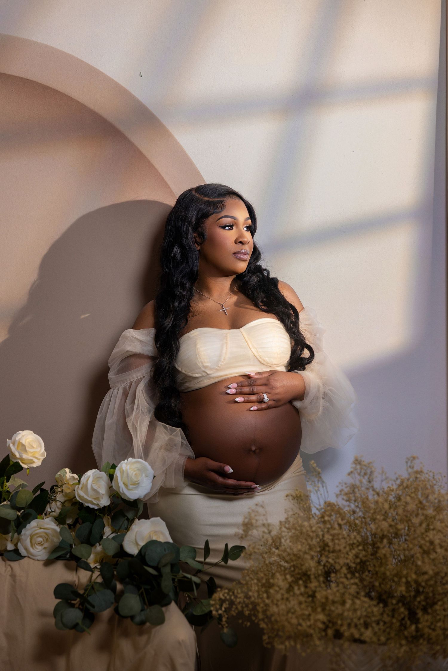 Maternity photoshoot featuring an elegant white dress against a soft neutral background with white roses and dried flowers.