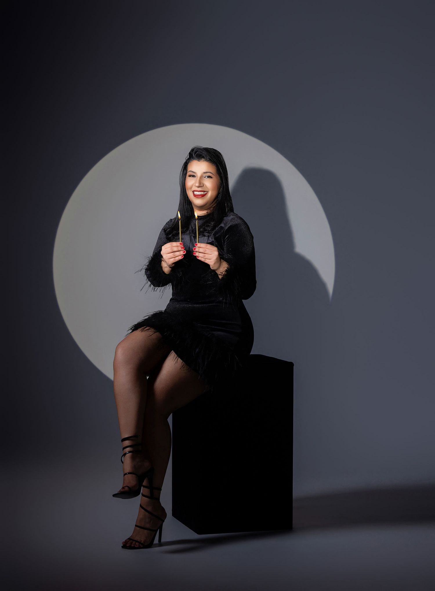 Woman in black velvet dress sitting on cube pedestal, smiling, holding sparklers against gray studio backdrop.