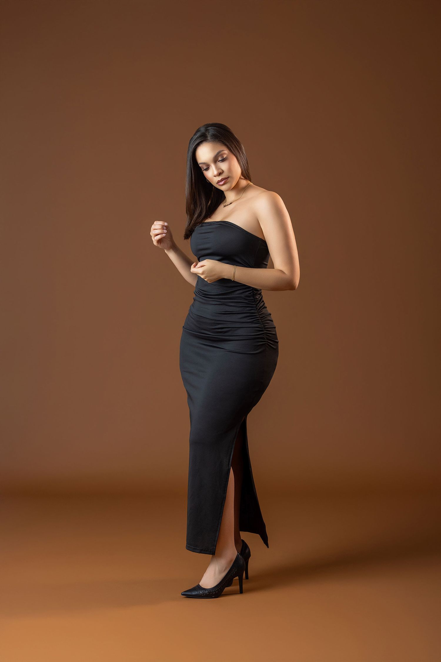 Woman in elegant black strapless ruched midi dress with slit poses in heels on brown background.