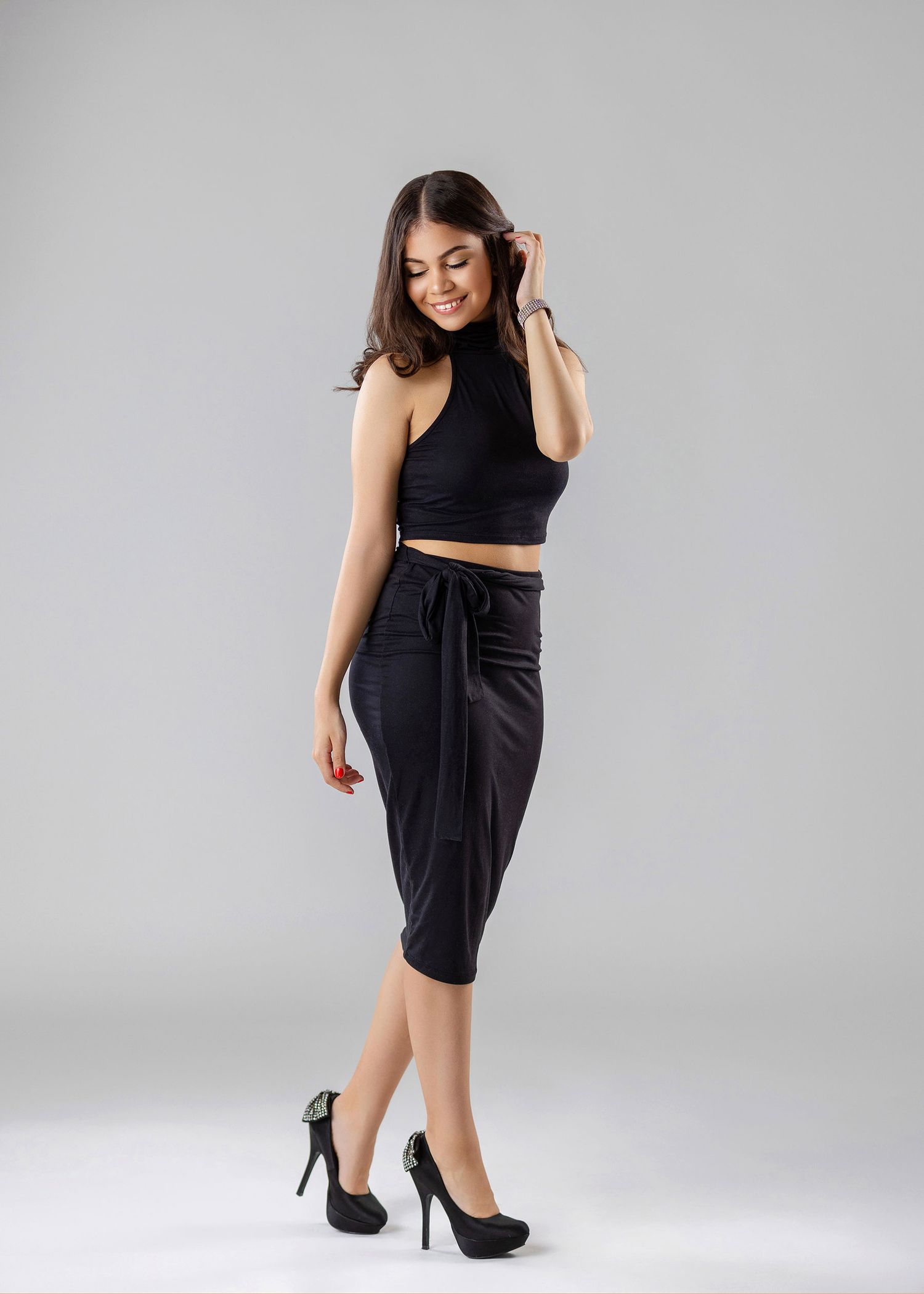 Woman in black halter crop top and midi skirt poses in high heels on gray background.