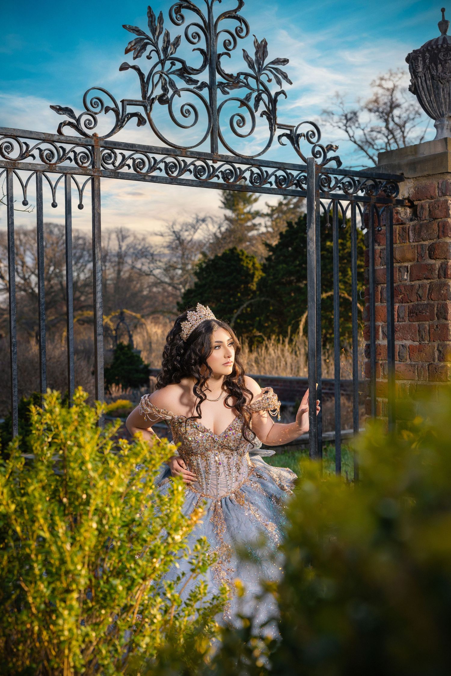 A young woman in a sparkling ball gown and tiara poses gracefully by ornate iron gates in a garden setting.