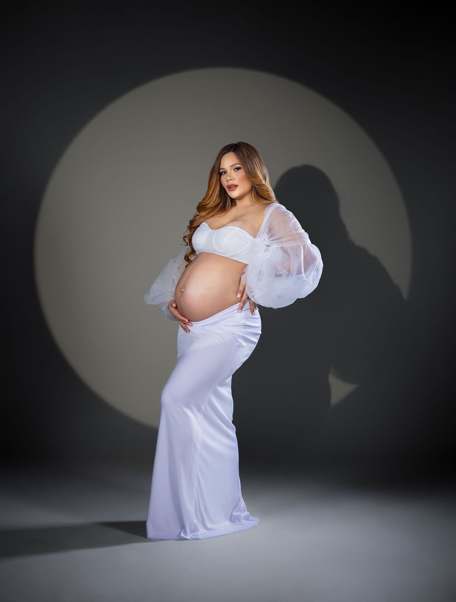 Artistic maternity photoshoot series in white dress against dark background showing pregnancy silhouette.
