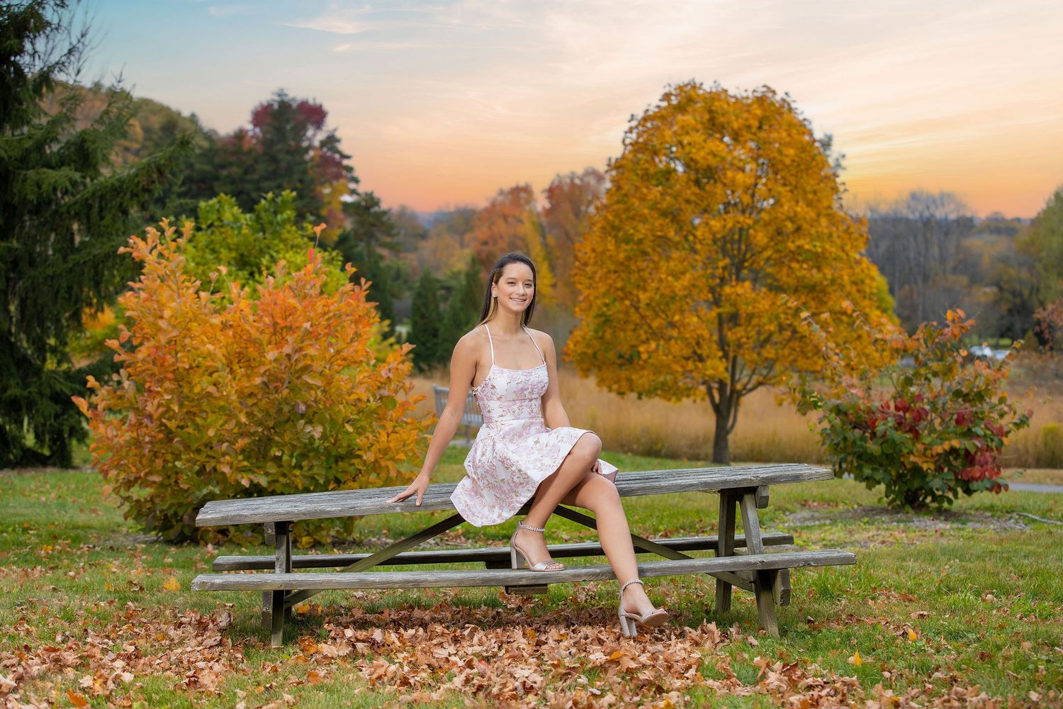 A white dress-clad figure sits on a wooden picnic table surrounded by vibrant autumn foliage at sunset.