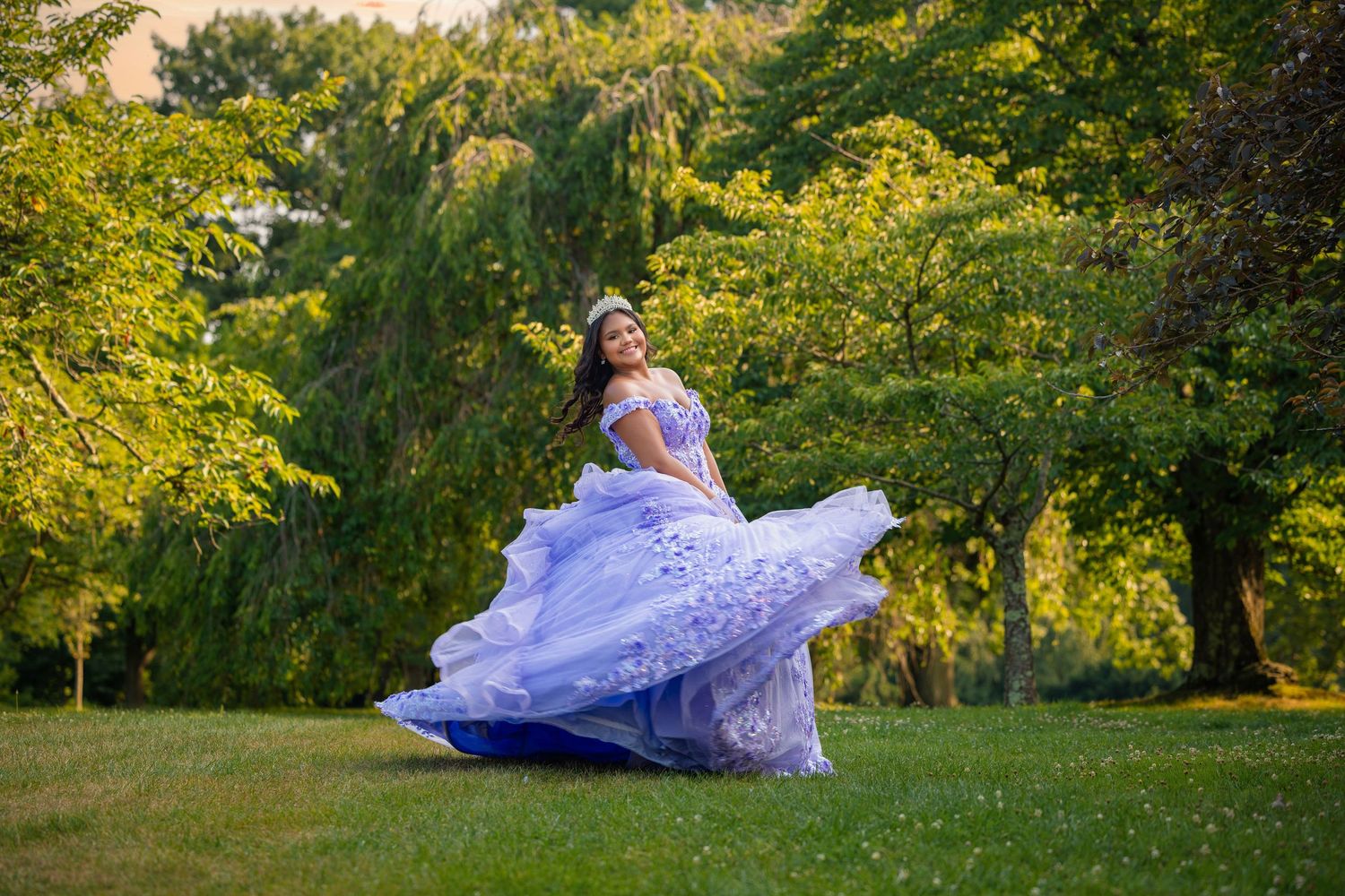 Young woman in flowing lavender quinceañera gown twirling gracefully in a lush green park during golden hour.