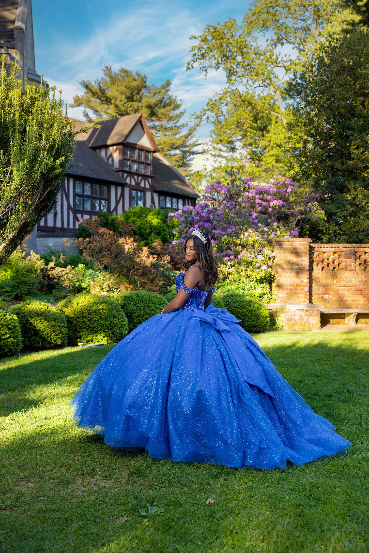 A vibrant royal blue ballgown dress flows across a sunlit garden lawn with Tudor-style architecture in the background.