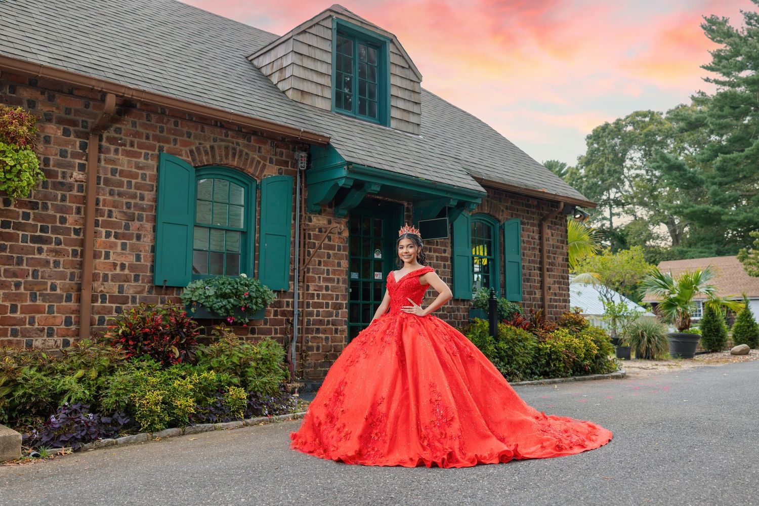 Young woman in a stunning red ball gown poses in front of a brick building at sunset.