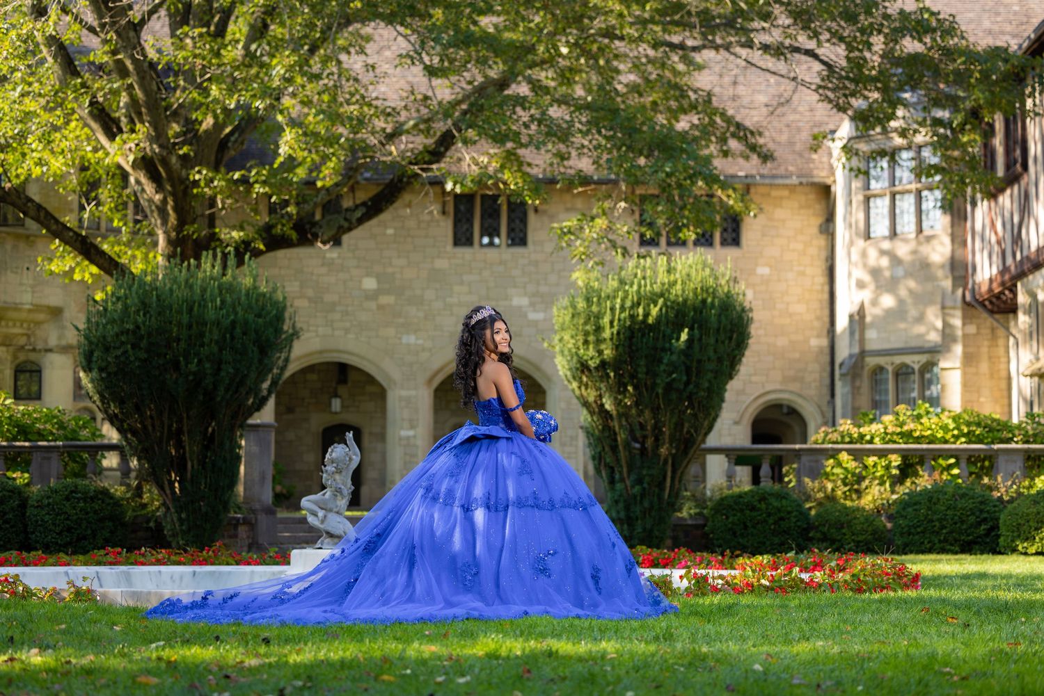 A person in a flowing royal blue quinceañera gown poses on a lawn in front of a stone mansion.