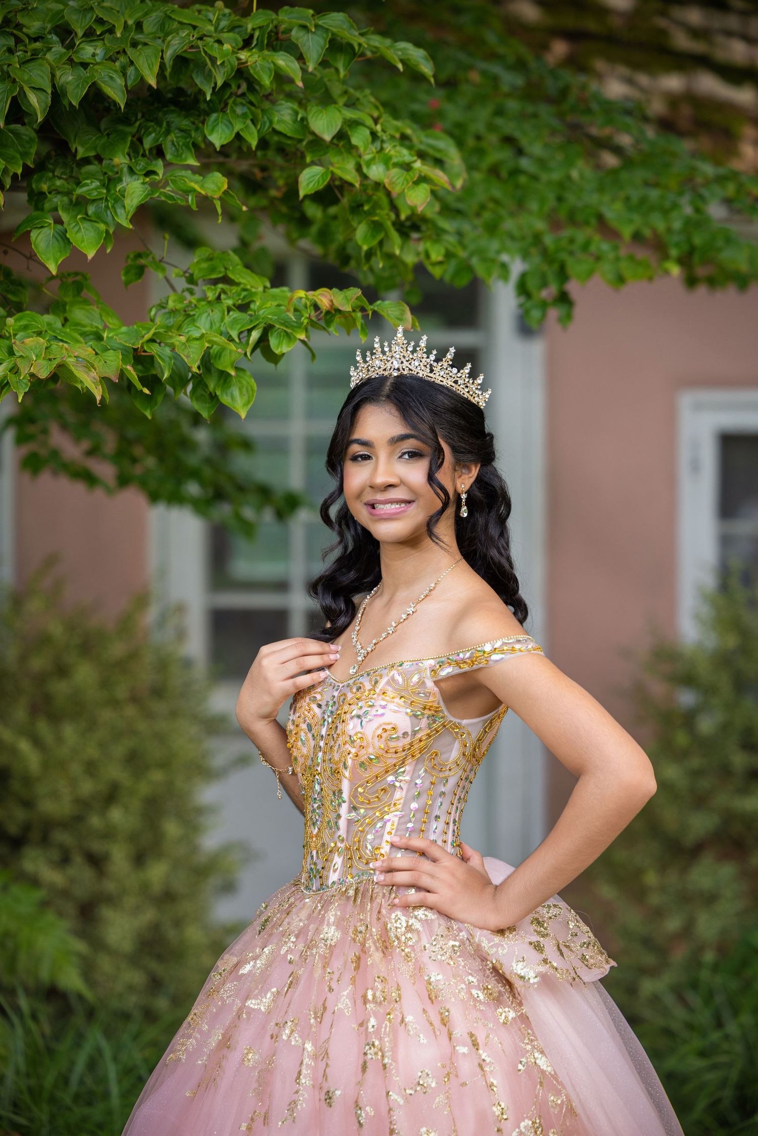 Young Latina woman in a pink and gold quinceañera ball gown and tiara crown posing outdoors.