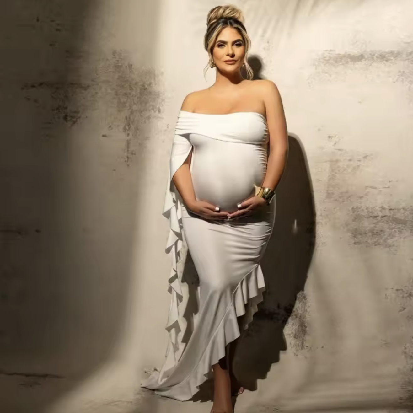 Pregnant woman in white off-shoulder ruffle maternity gown posing gracefully against a neutral studio backdrop.