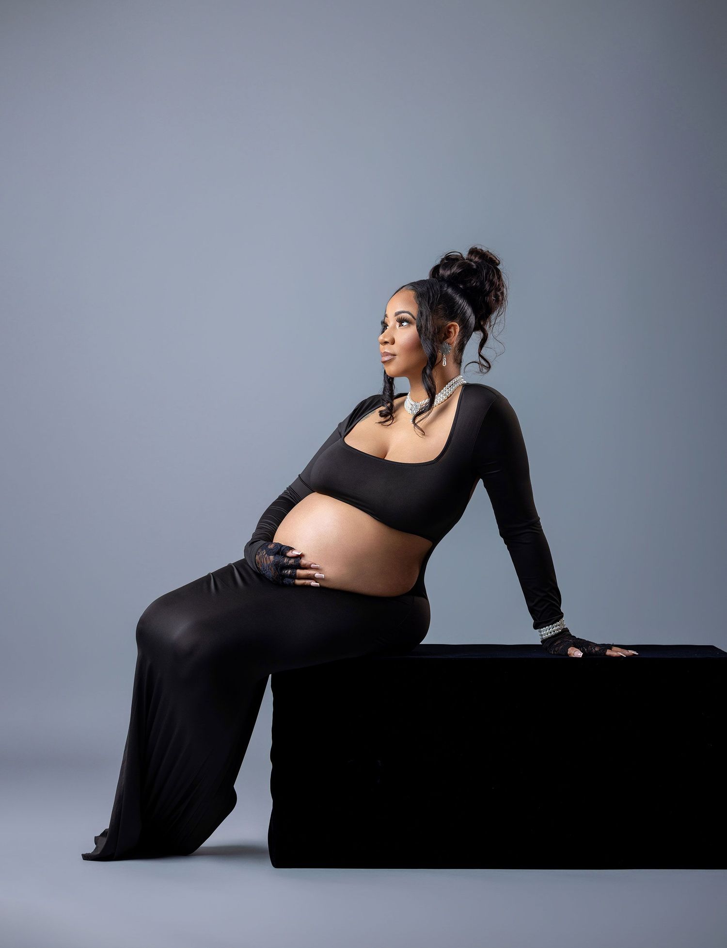 Silhouette of a pregnant woman in a black dress reclining on a dark surface against a gray studio backdrop.