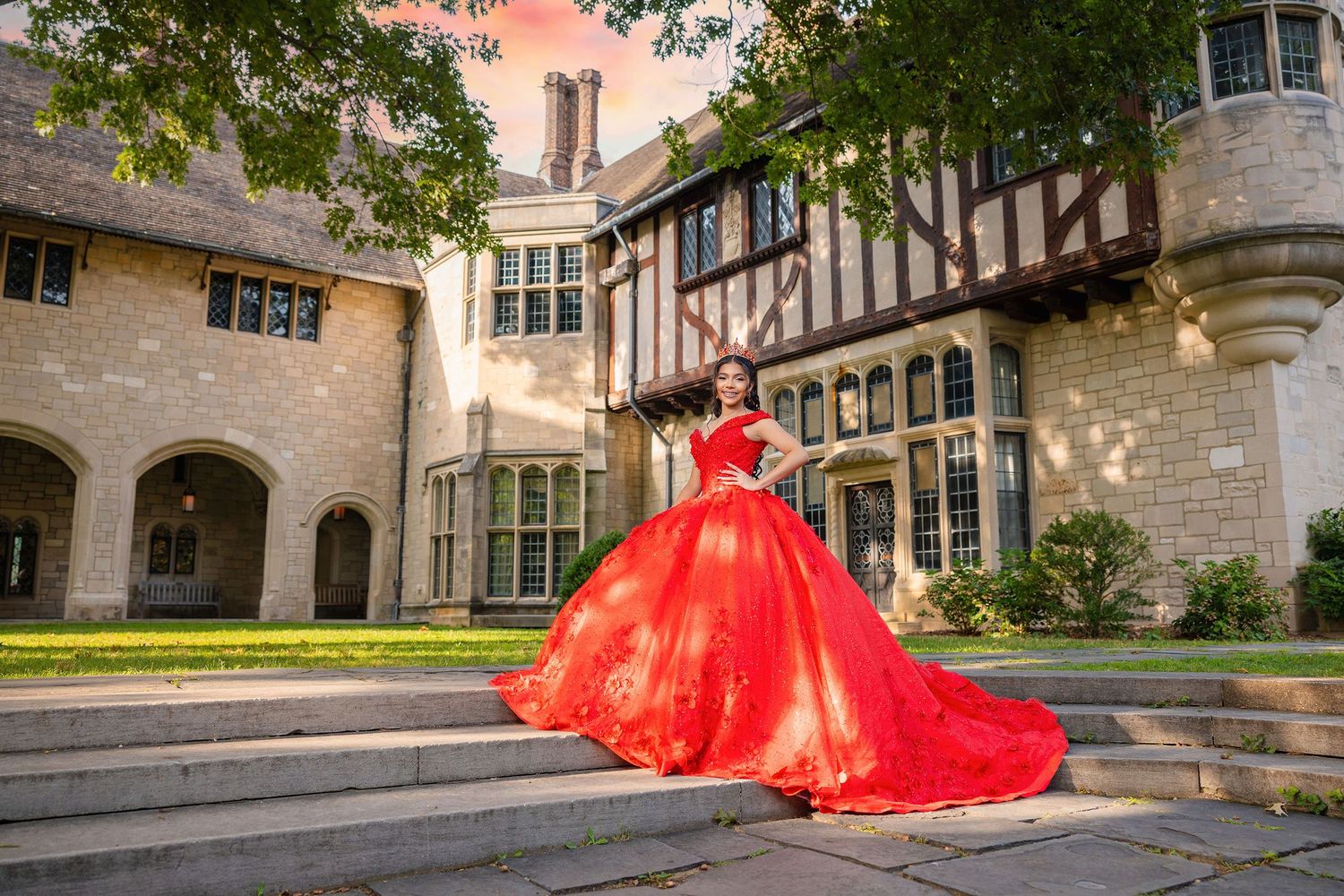A dramatic red quinceañera dress photographed in front of a grand stone building with Tudor-style architecture.