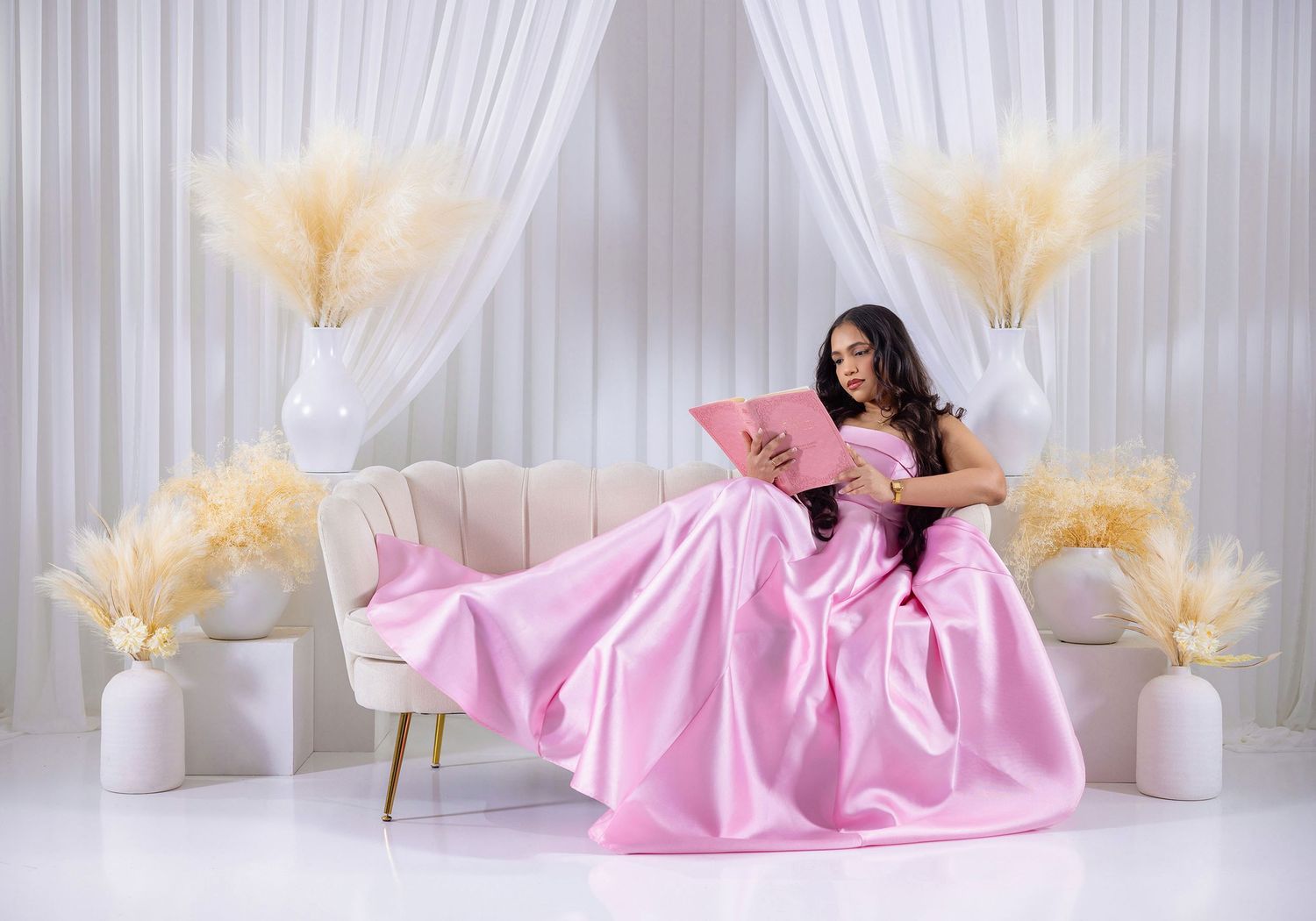 Woman in elegant pink ball gown reclining on cream sofa with pampas grass decor.