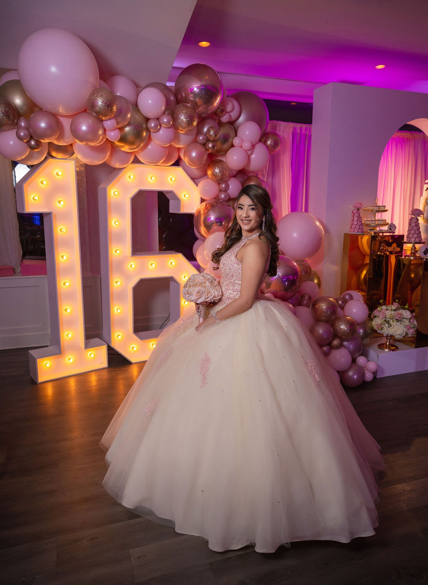 Elegant quinceañera celebration with pink lighting, large illuminated letters and balloon decor in an indoor venue.