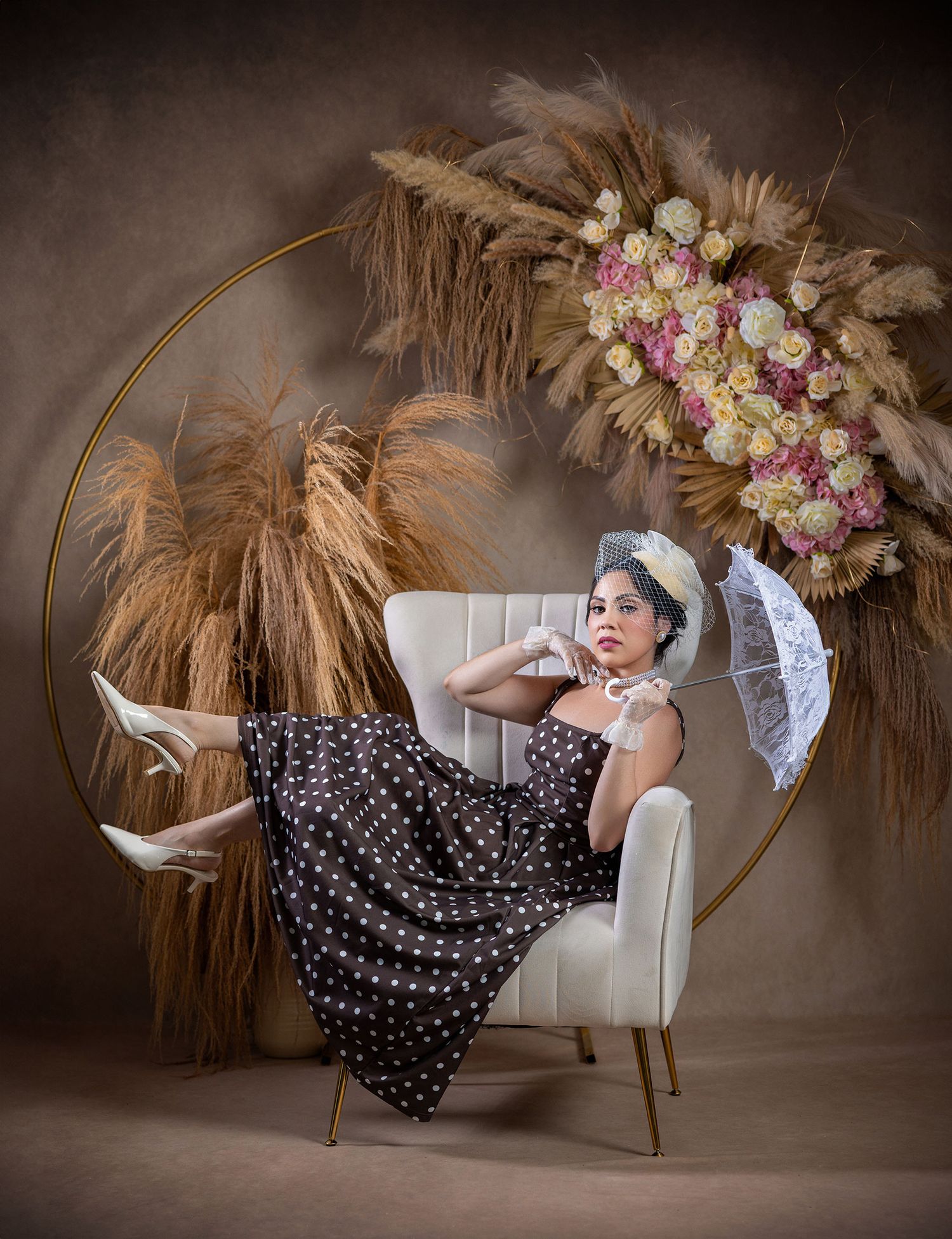 Woman in polka dot dress poses in chair with floral hoop backdrop and parasol.