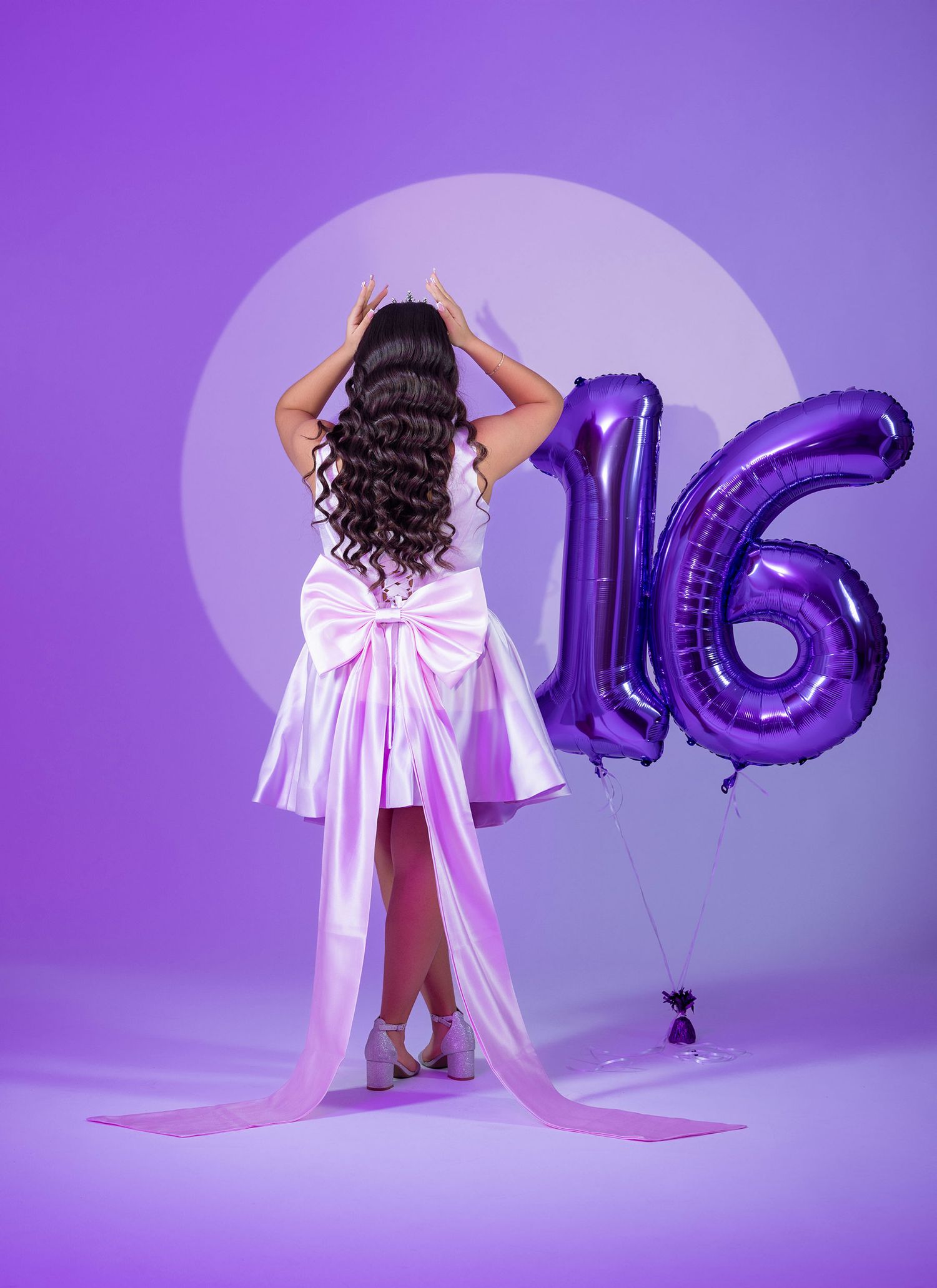 Teen in white dress with large bow standing beside purple number 16 balloons on purple backdrop.