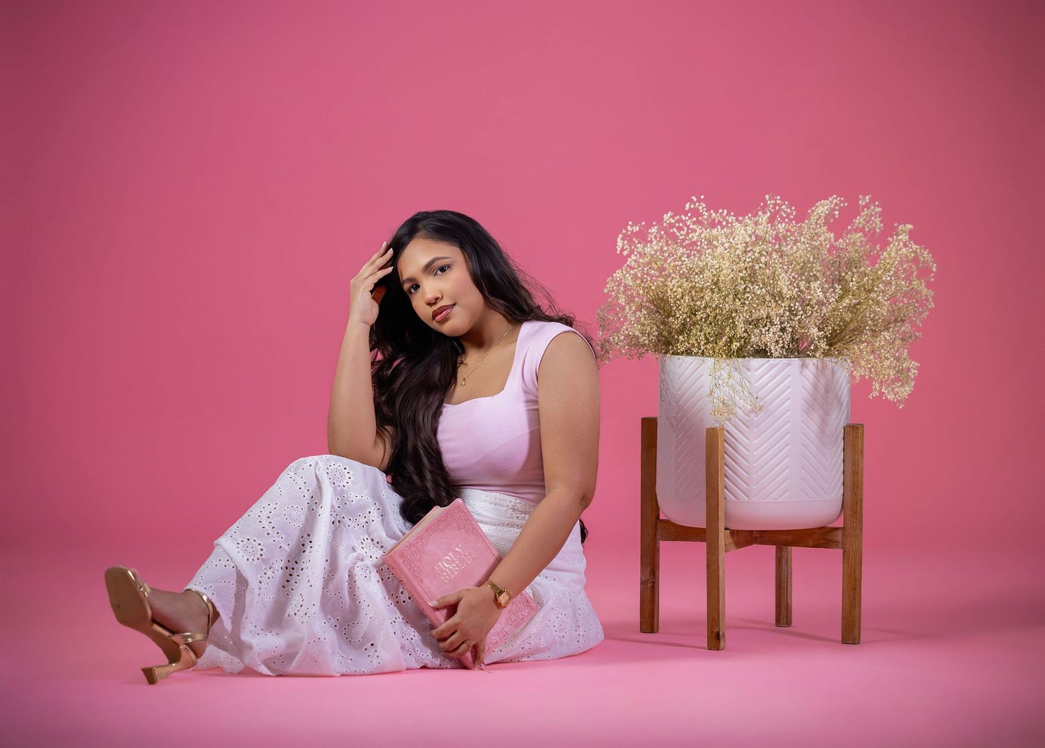Woman in white outfit sitting on floor beside pink floral arrangement on wooden stand, pink background.
