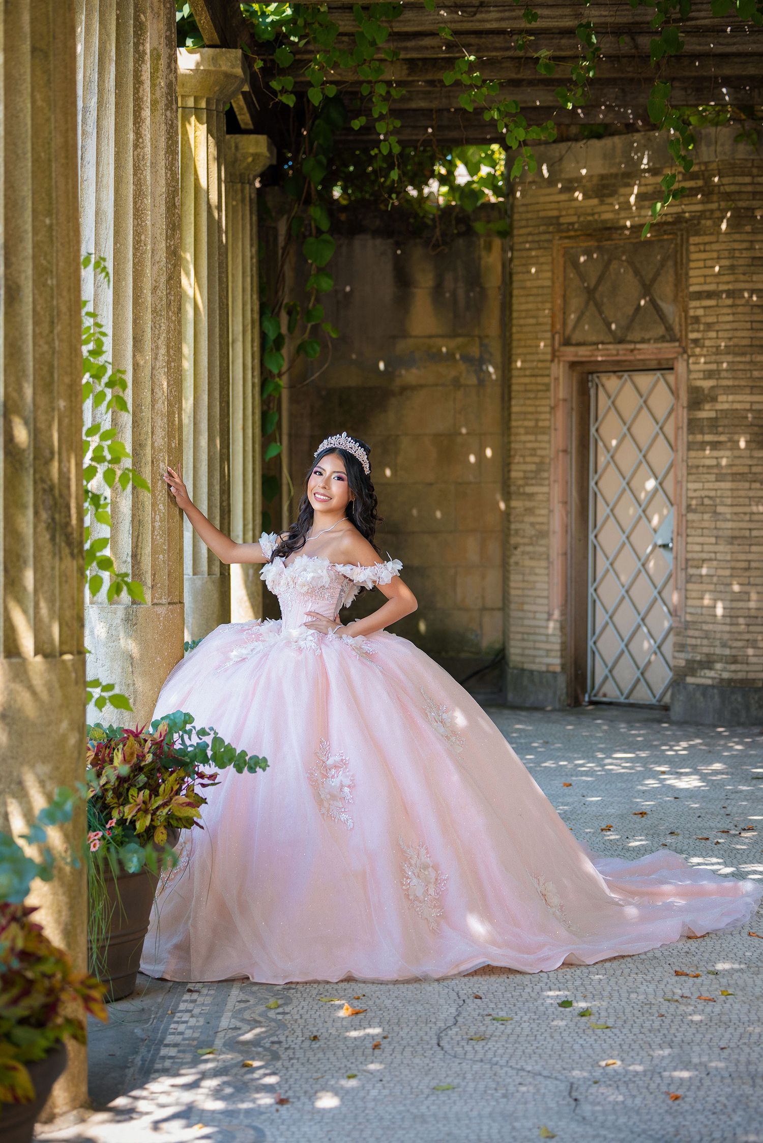 A stunning pink quinceañera ball gown photographed in an elegant outdoor courtyard setting with brick columns.
