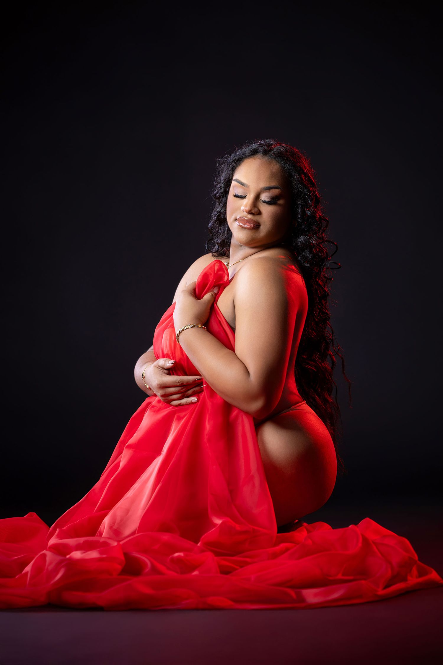 Pregnant woman in flowing red sheer fabric poses gracefully on studio floor against black background.