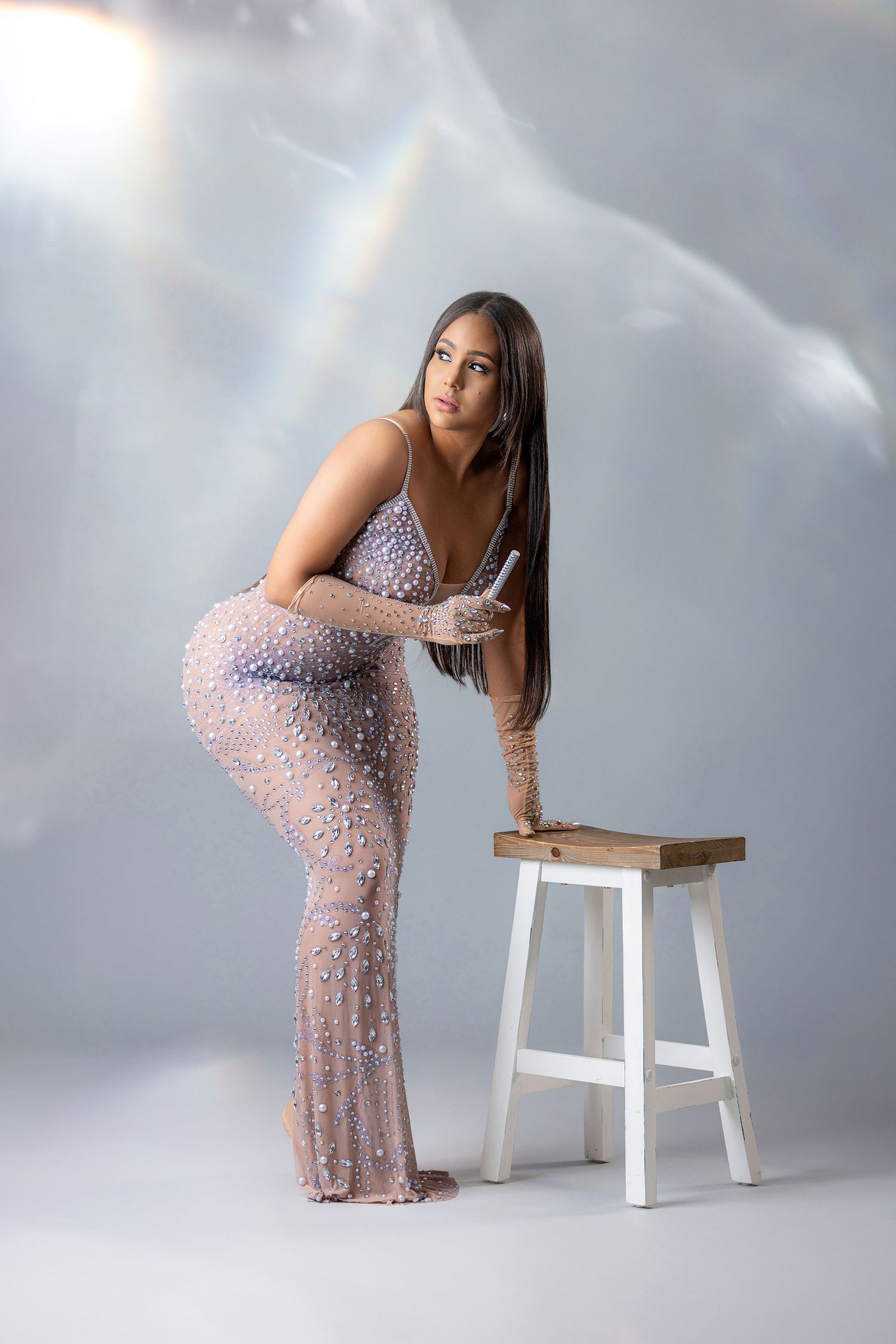 Woman in sheer beaded gown posing beside white wooden stool against misty gray studio backdrop.